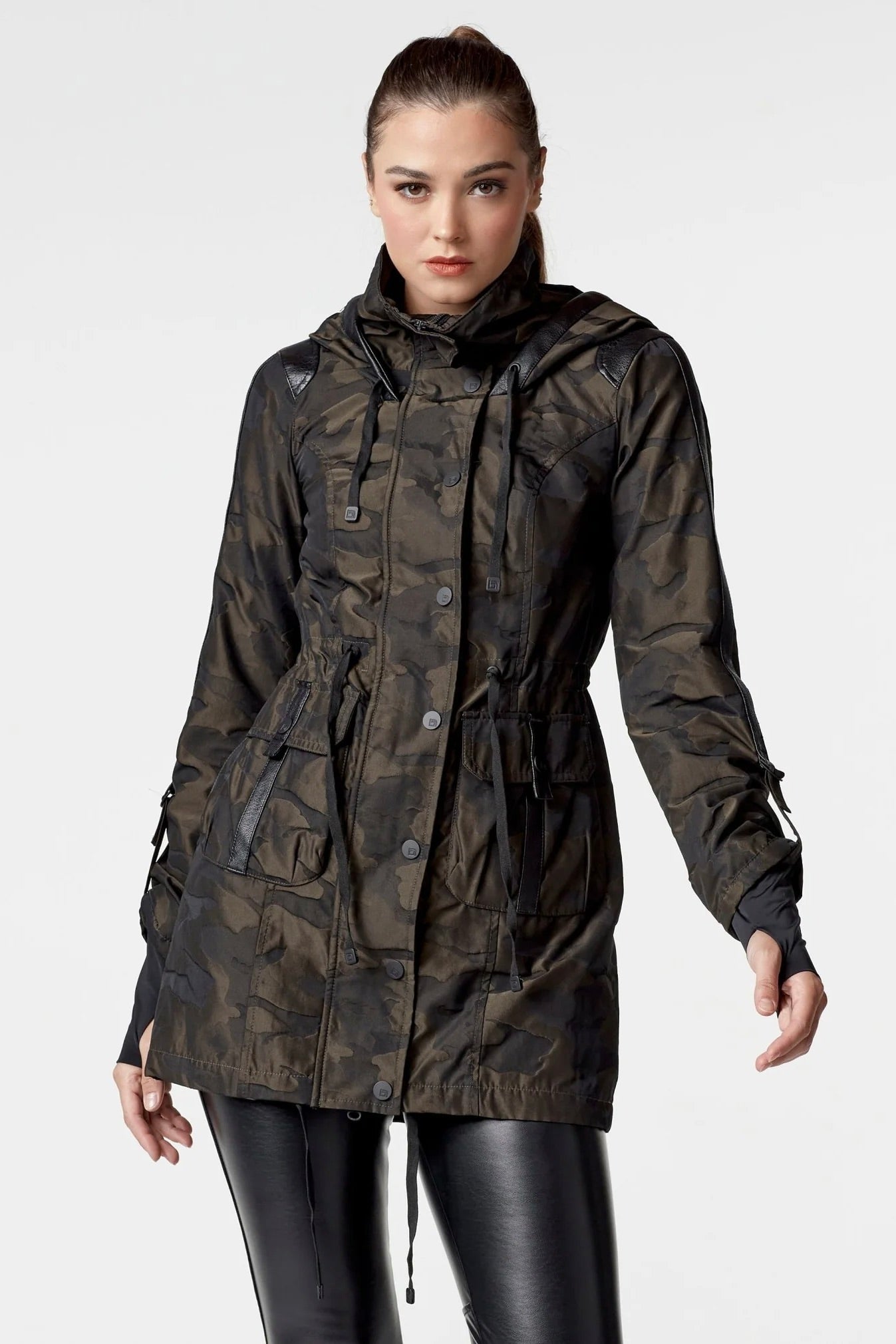 Woman wearing an olive camo anorak jacket with adjustable waist, hood, storm cuffs with thumbholes, zip front with button closure, and two front pockets. Stylish equestrian outerwear designed as a versatile horse riding jacket, barn jacket, and raincoat for horse girls.