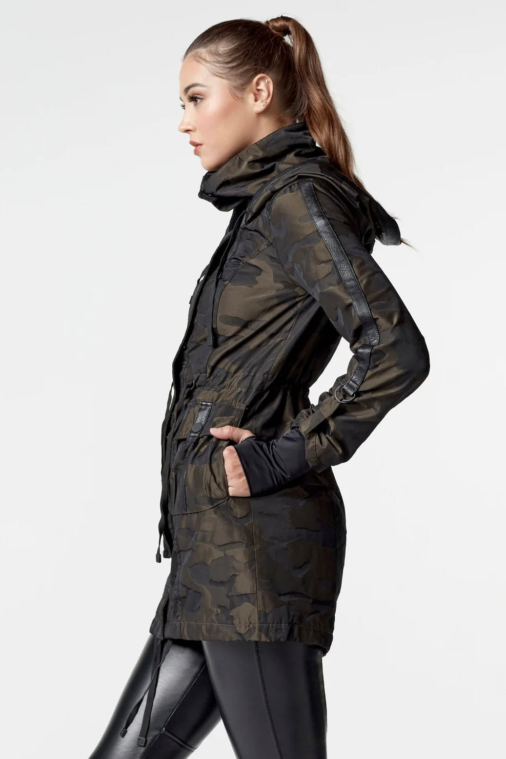 Woman wearing an olive camo anorak jacket with adjustable waist, hood, storm cuffs with thumbholes, zip front with button closure, and two front pockets. Stylish equestrian outerwear designed as a versatile horse riding jacket, barn jacket, and raincoat for horse girls.