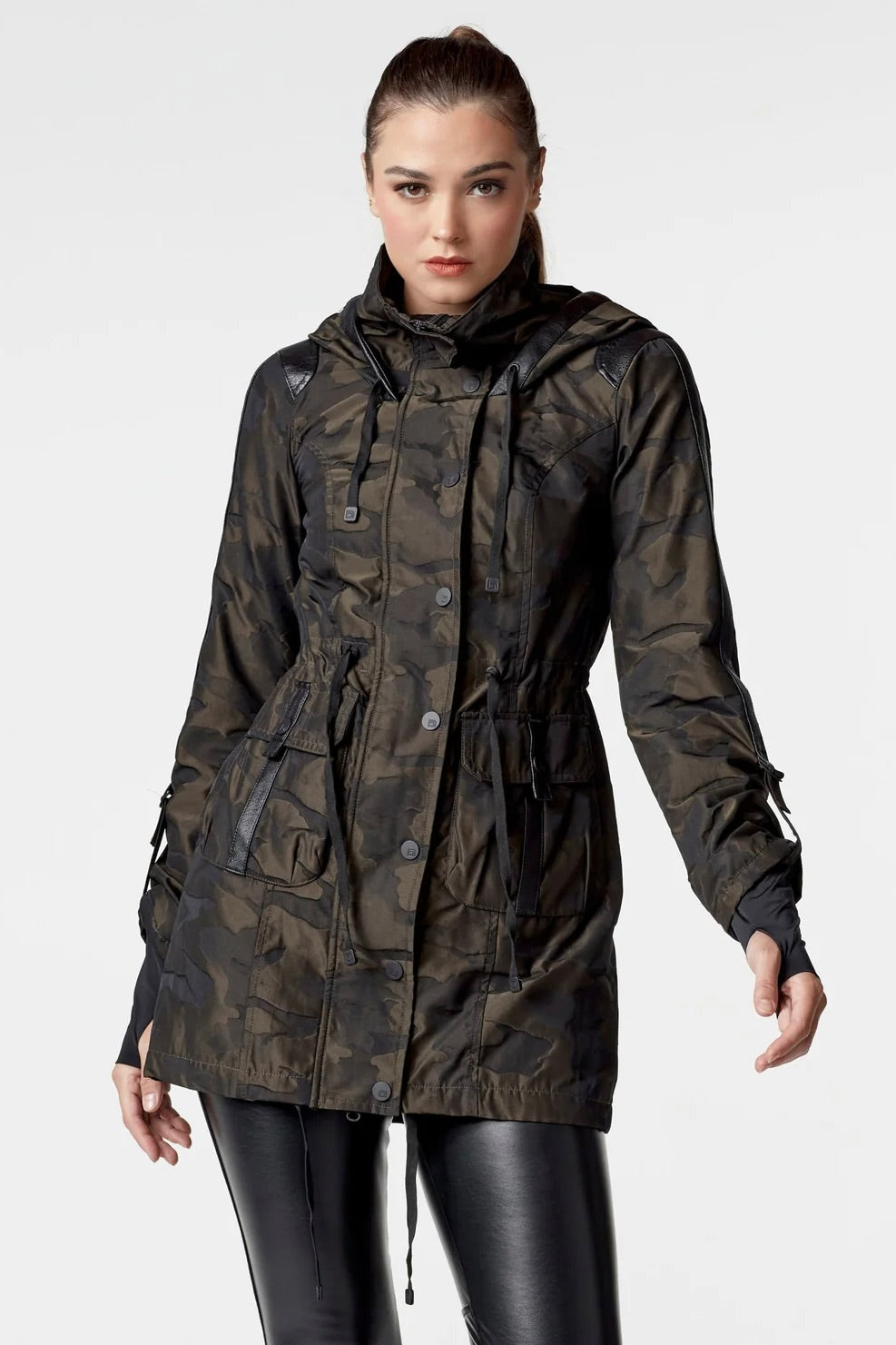 Woman wearing an olive camo anorak jacket with adjustable waist, hood, storm cuffs with thumbholes, zip front with button closure, and two front pockets. Stylish equestrian outerwear designed as a versatile horse riding jacket, barn jacket, and raincoat for horse girls.