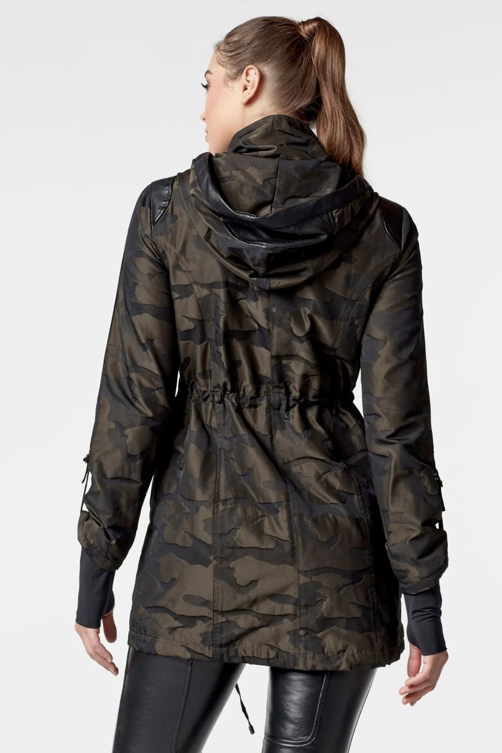Woman wearing an olive camo anorak jacket with adjustable waist, hood, storm cuffs with thumbholes, zip front with button closure, and two front pockets. Stylish equestrian outerwear designed as a versatile horse riding jacket, barn jacket, and raincoat for horse girls.