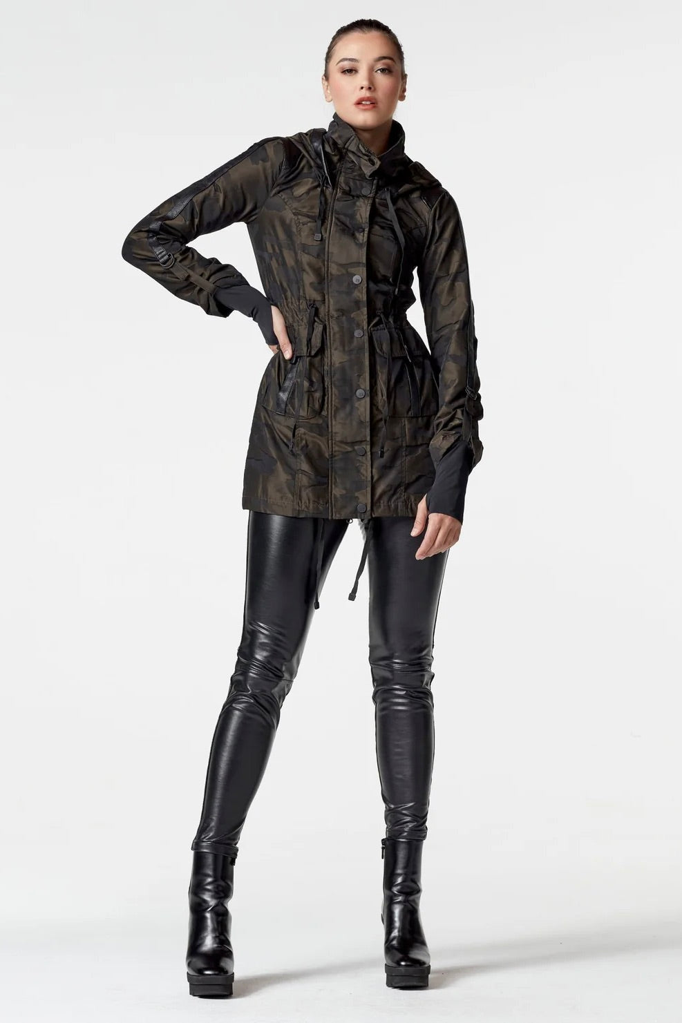 Woman wearing an olive camo anorak jacket with adjustable waist, hood, storm cuffs with thumbholes, zip front with button closure, and two front pockets. Stylish equestrian outerwear designed as a versatile horse riding jacket, barn jacket, and raincoat for horse girls.