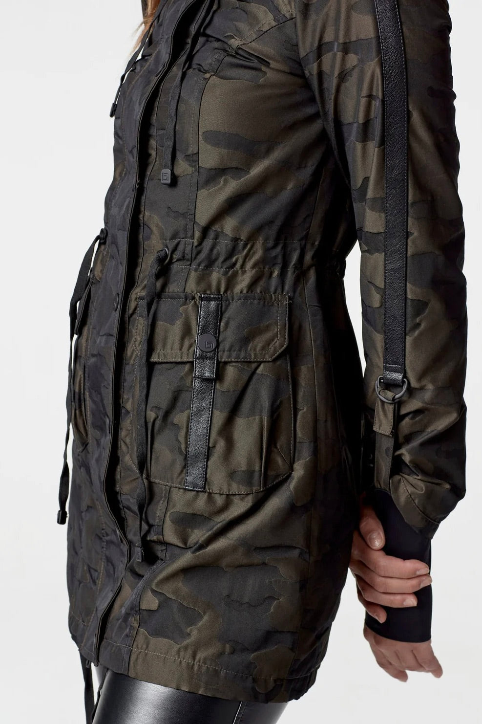 Woman wearing an olive camo anorak jacket with adjustable waist, hood, storm cuffs with thumbholes, zip front with button closure, and two front pockets. Stylish equestrian outerwear designed as a versatile horse riding jacket, barn jacket, and raincoat for horse girls.