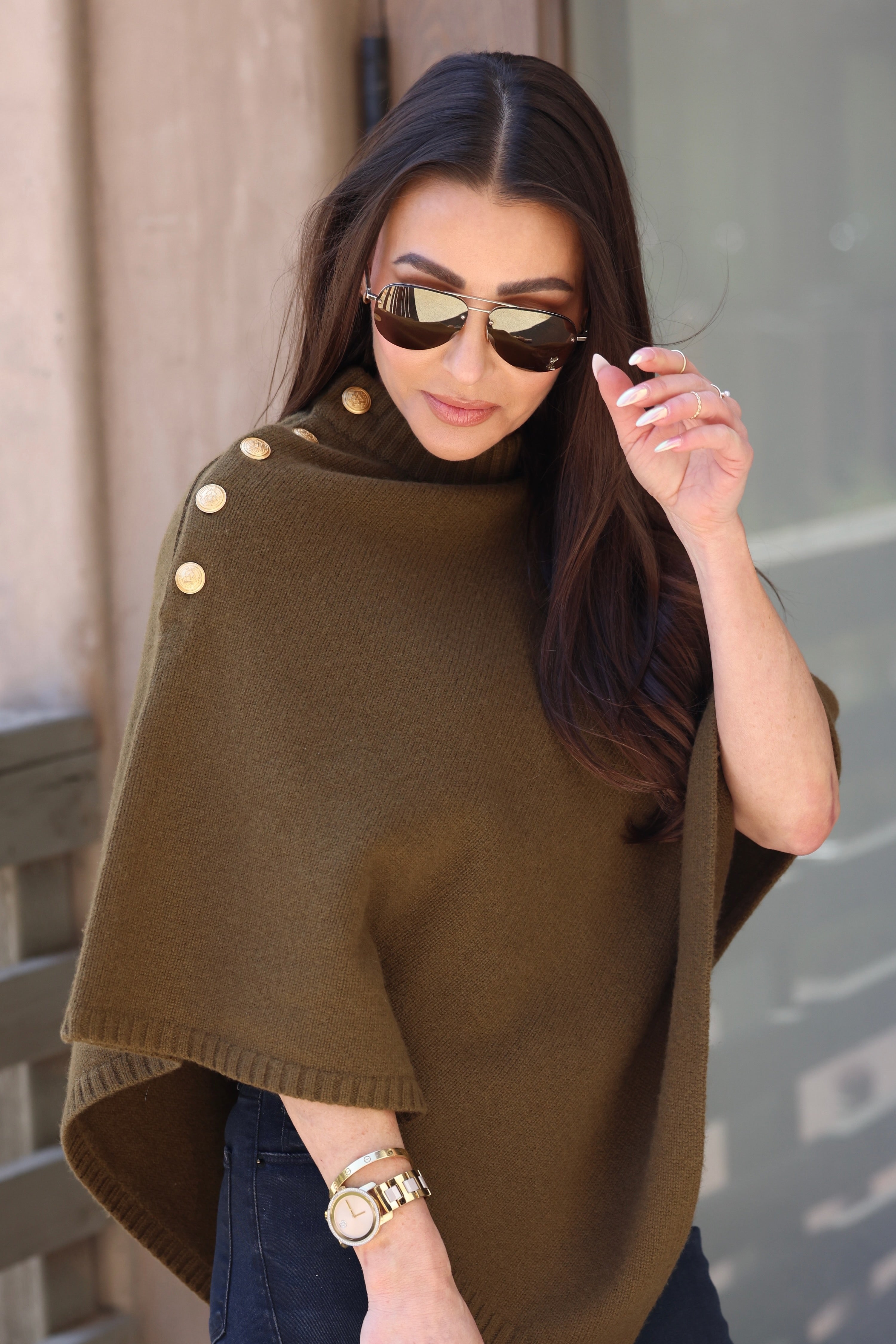 Woman wearing sunglasses and an olive poncho with gold buttons, adjusting her hair. She has long dark hair, minimal makeup, and a gold watch.