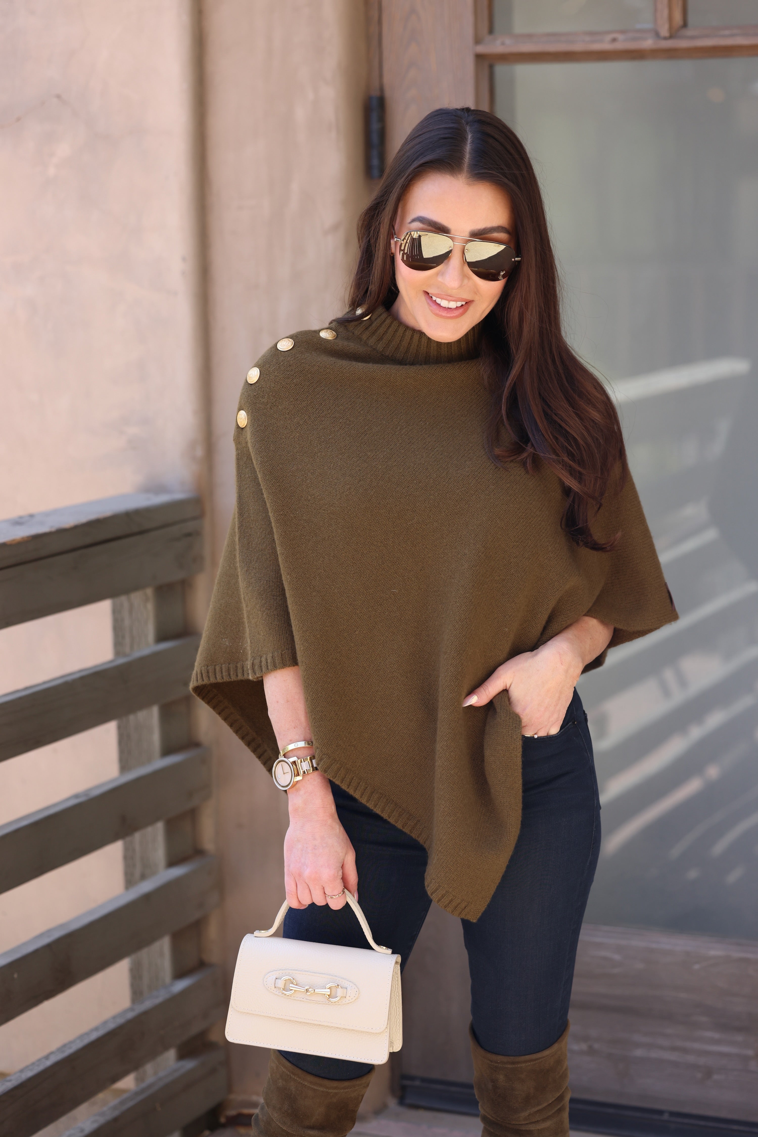 A woman in sunglasses smiles, wearing an olive poncho with gold buttons. She holds a small beige handbag and stands confidently near a wooden railing.