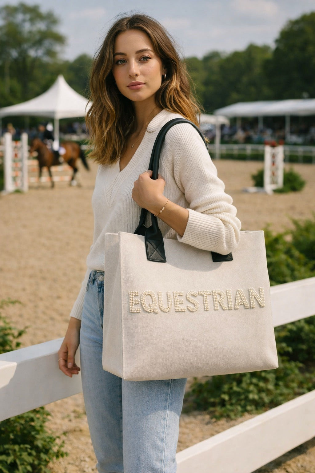 Woman holding a beige tote bag with 'Equestrian' text in an outdoor setting with horses.