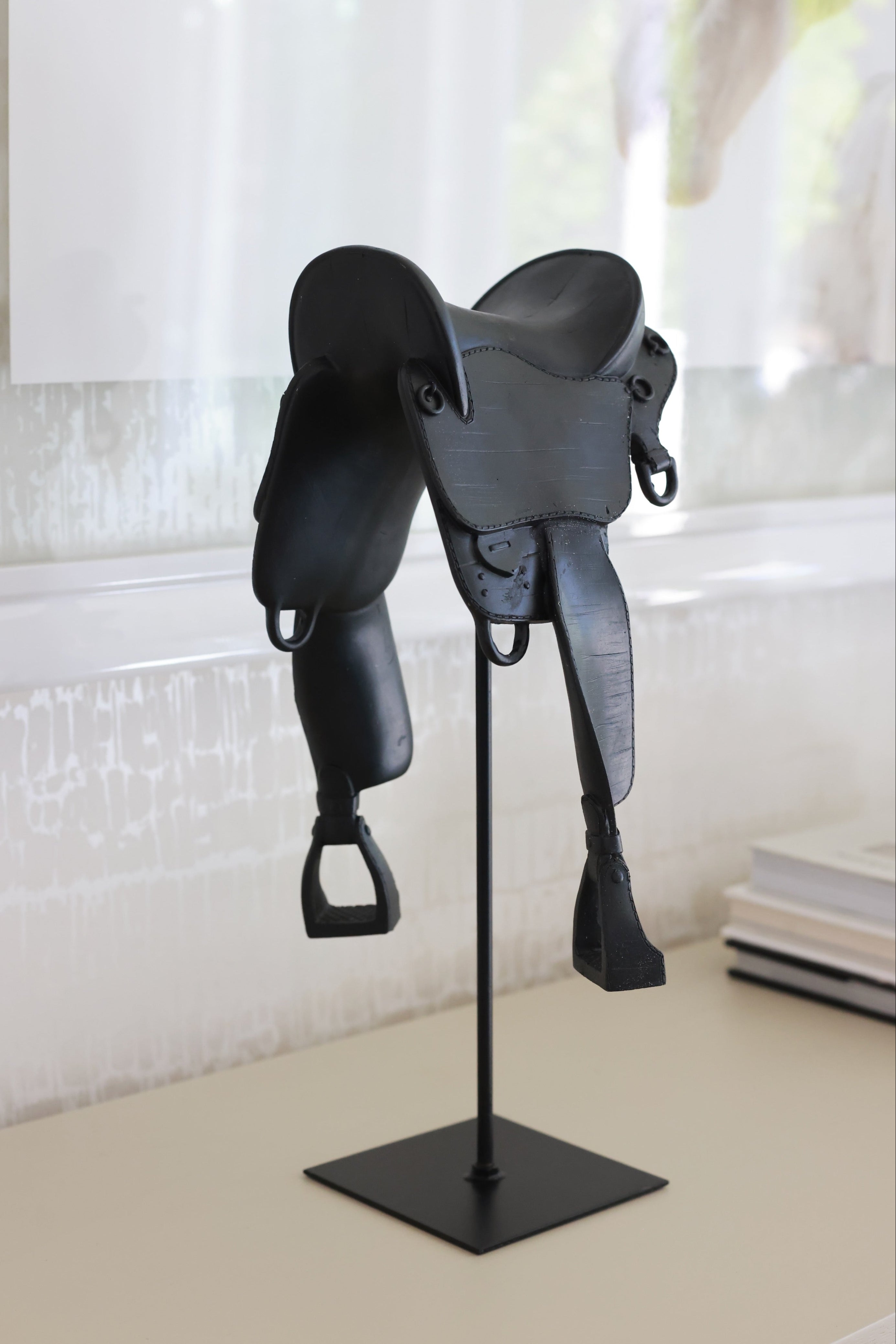 Black English-style horse saddle decor piece crafted from matte-finish resin and metal detailing. Mounted on a black metal stand with a square base, this modern equestrian sculpture captures the elegance of traditional riding saddles and adds a bold statement to home or office décor.