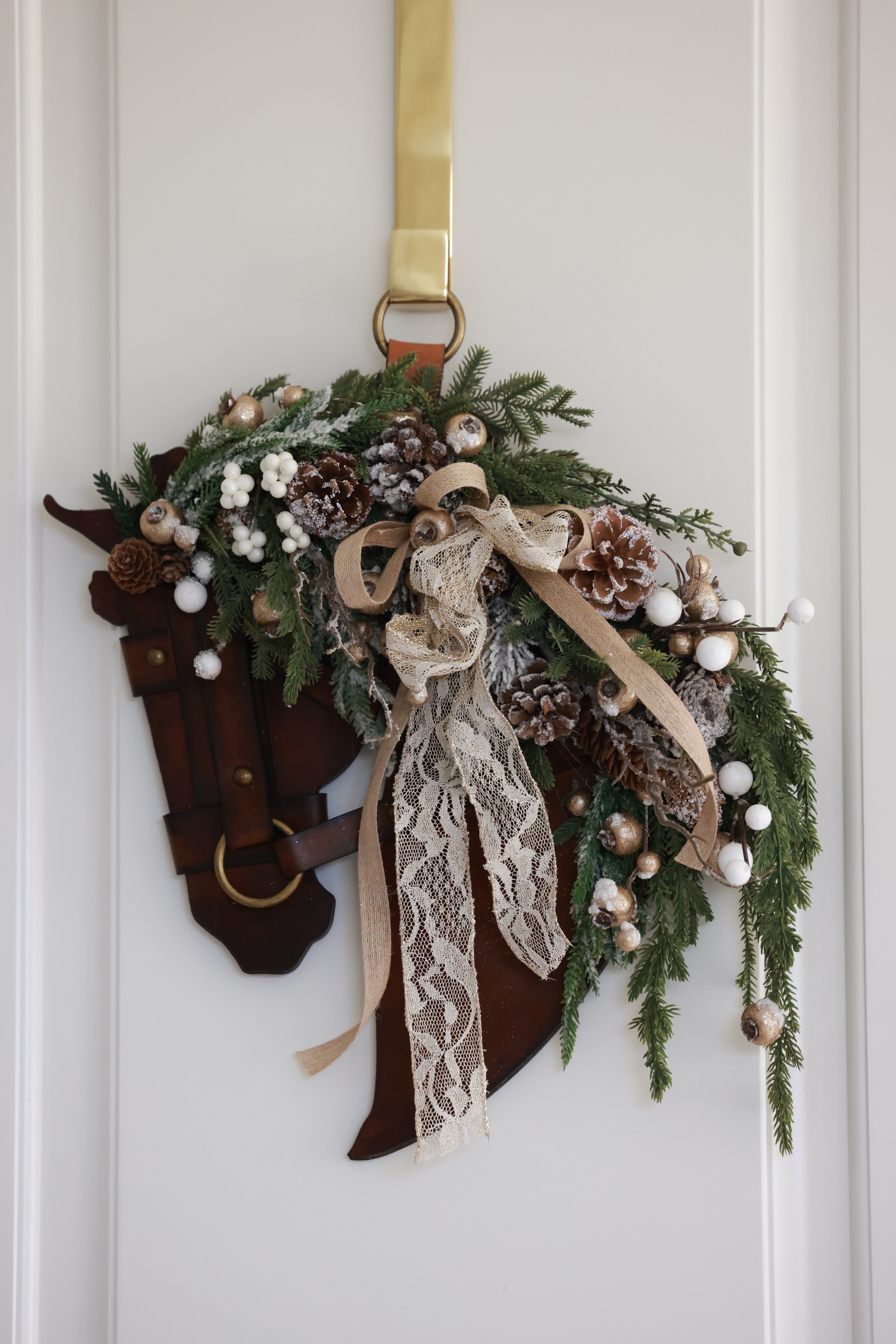 A handcrafted equestrian holiday wreath featuring a sculpted genuine leather horse profile adorned with winter greenery, frosted pinecones, pearl-accented berries, shimmering metallic details, and a layered lace and linen bow. The wreath measures approximately 16 inches by 12 inches and is designed for indoor or outdoor display.