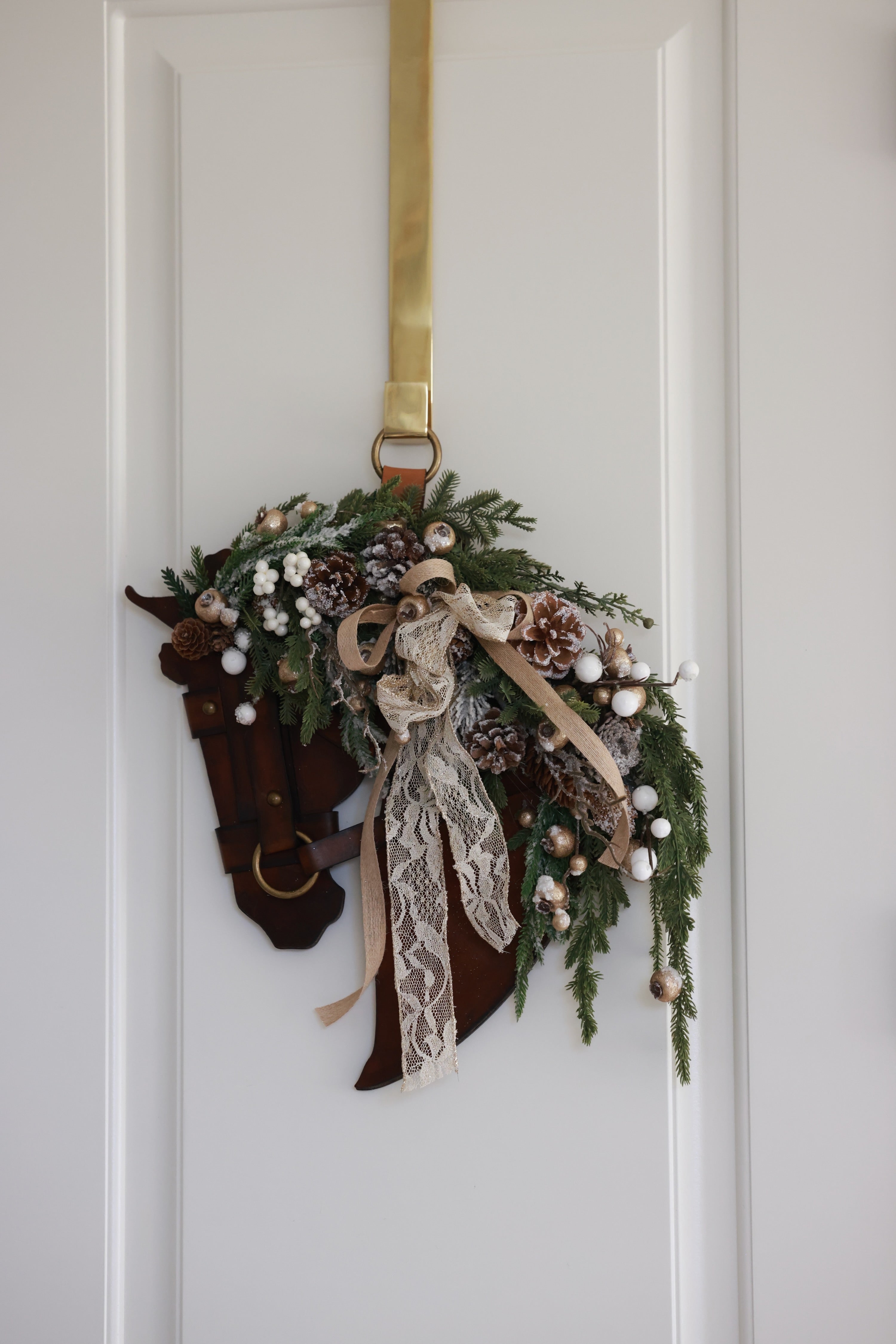 A handcrafted equestrian holiday wreath featuring a sculpted genuine leather horse profile adorned with winter greenery, frosted pinecones, pearl-accented berries, shimmering metallic details, and a layered lace and linen bow. The wreath measures approximately 16 inches by 12 inches and is designed for indoor or outdoor display.