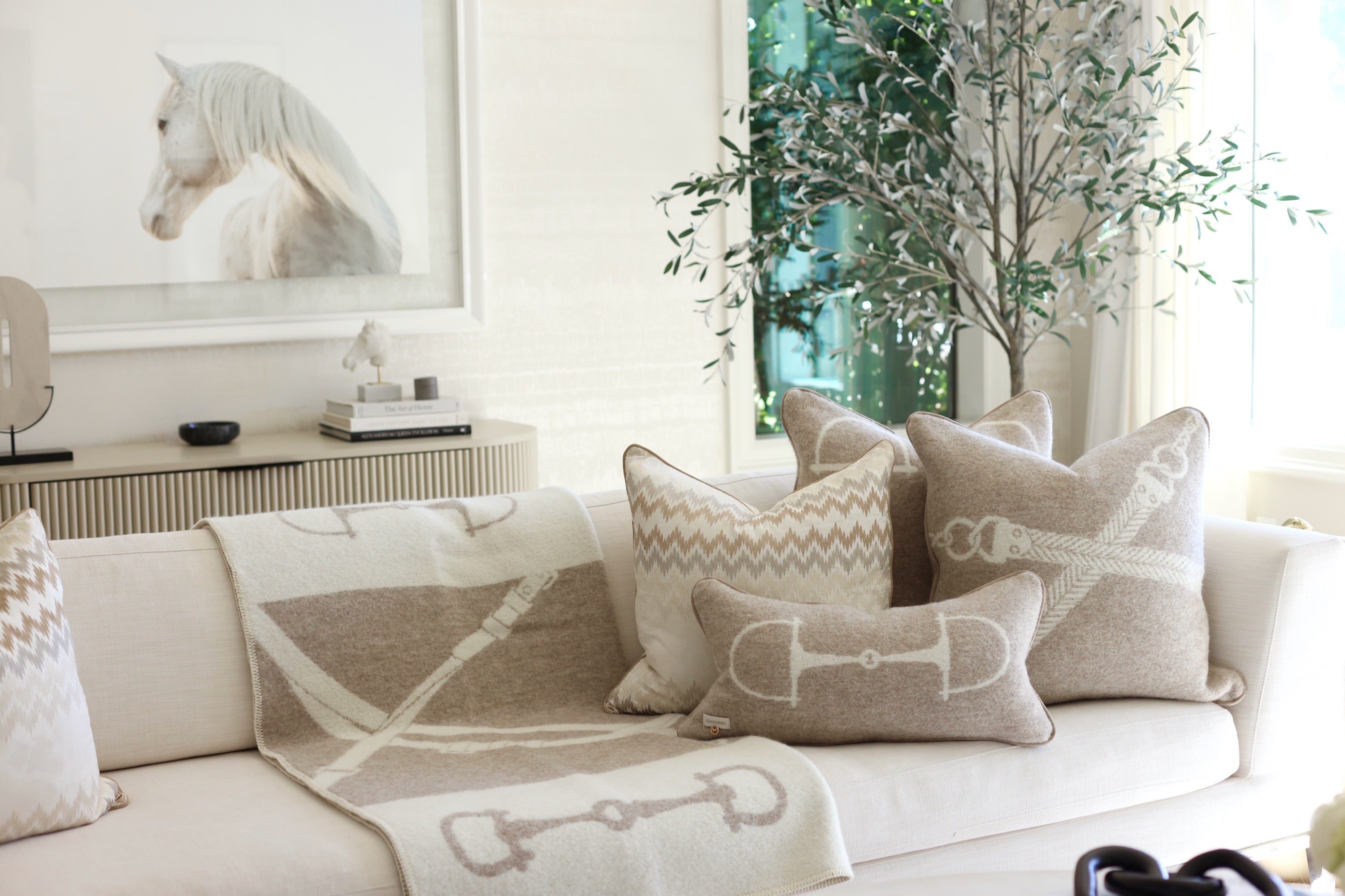 Equine Lumbar Wool Pillow - Vanilla Bean A cozy living room scene with a white sofa adorned with beige and white patterned pillows and a matching blanket. A green indoor tree stands beside it. Luxury rectangular cushion in vanilla bean, made from 100% New Zealand lambswool with ADAMSBRO’s signature equestrian bit pattern. Handcrafted in Sweden, featuring faux suede piping, concealed zipper, and a plush duck feather insert, a refined accent for timeless, Scandinavian-inspired interiors.