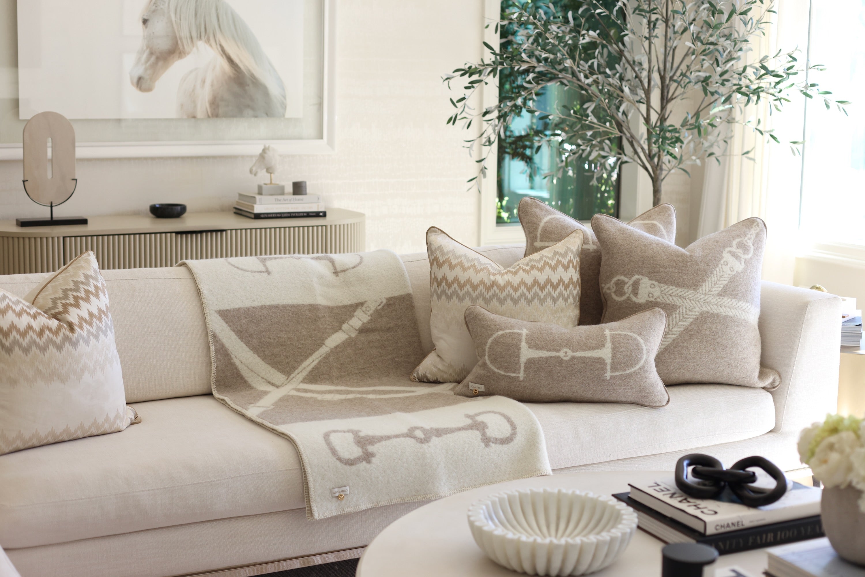 Equine Wool Throw - Vanilla Bean | A cozy living room features a beige sofa with a horse-themed blanket and pillow. A framed horse photo and potted plant add a serene touch. Classic equestrian-inspired wool throw blanket in vanilla bean, crafted from 100% New Zealand lambswool. Features a reversible horse bit pattern, soft and breathable texture, and Scandinavian design, perfect for layering on sofas, beds, or armchairs in timeless, natural interiors