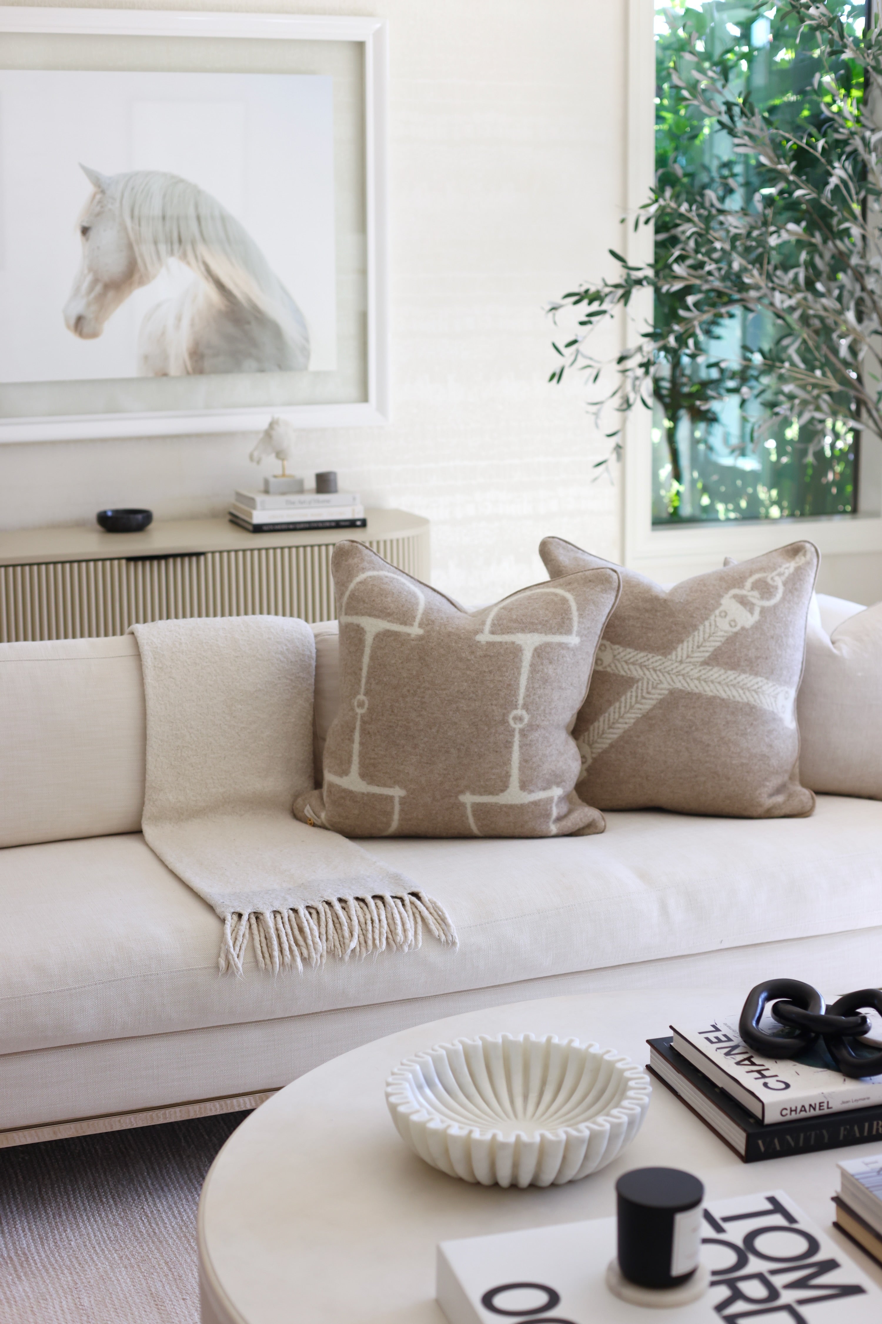 Adamsbro Equine Wool Pillow - Vanilla Bean | Cozy beige sofa with two horse bit patterned pillows; soft texture and neutral tones. A potted plant and framed art create a serene, minimalist ambiance. Luxury cushion in 100 new zealand lambswool | Luxury rectangular cushion handcrafted in Sweden from 100% New Zealand lambswool featuring an equestrian pattern, soft faux suede piping, and filled with plush duck feathers, a Scandinavian-inspired accent pillow