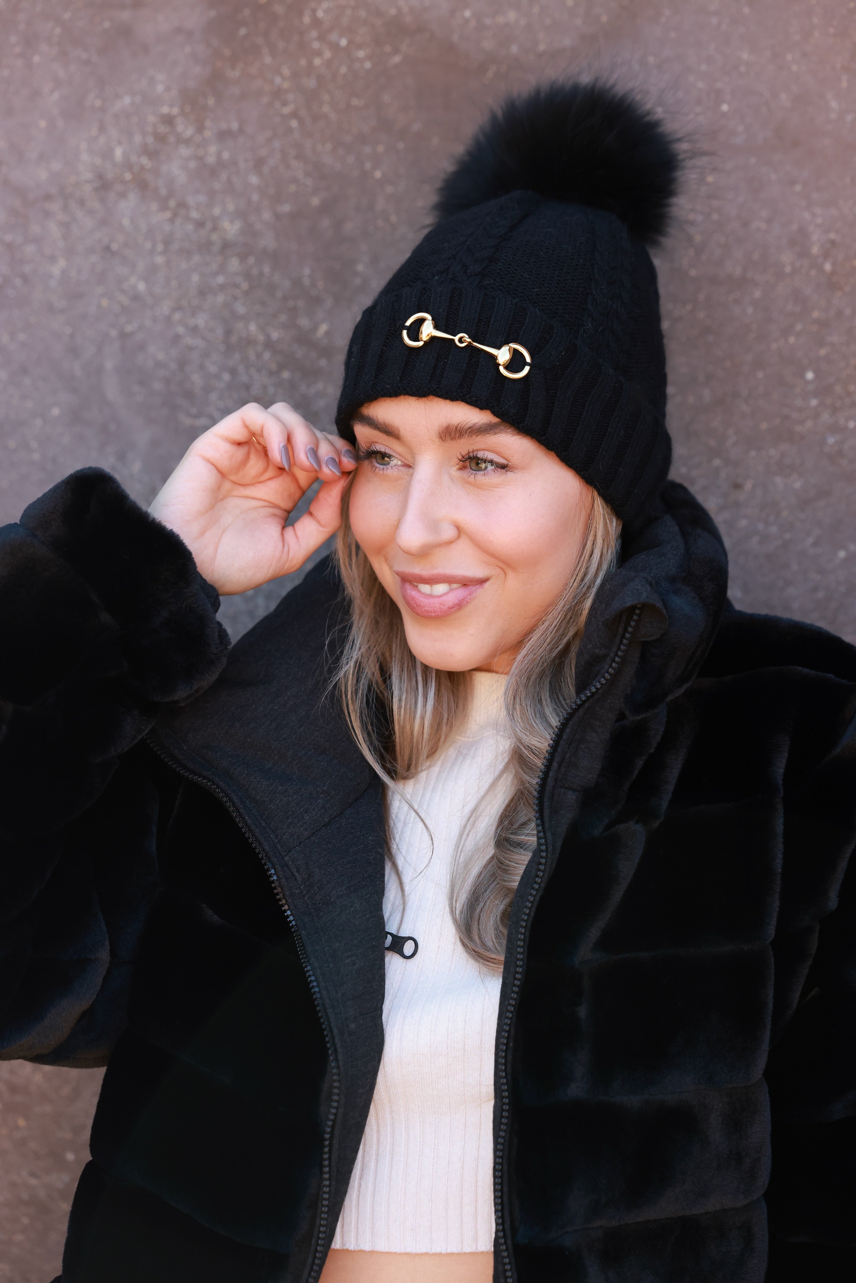 Black Evie Beanie featuring soft, cozy medium-knit cable design with polished gold bit accent and removable faux fur pom. A stylish equestrian winter accessory that adds a chic touch to cold days.