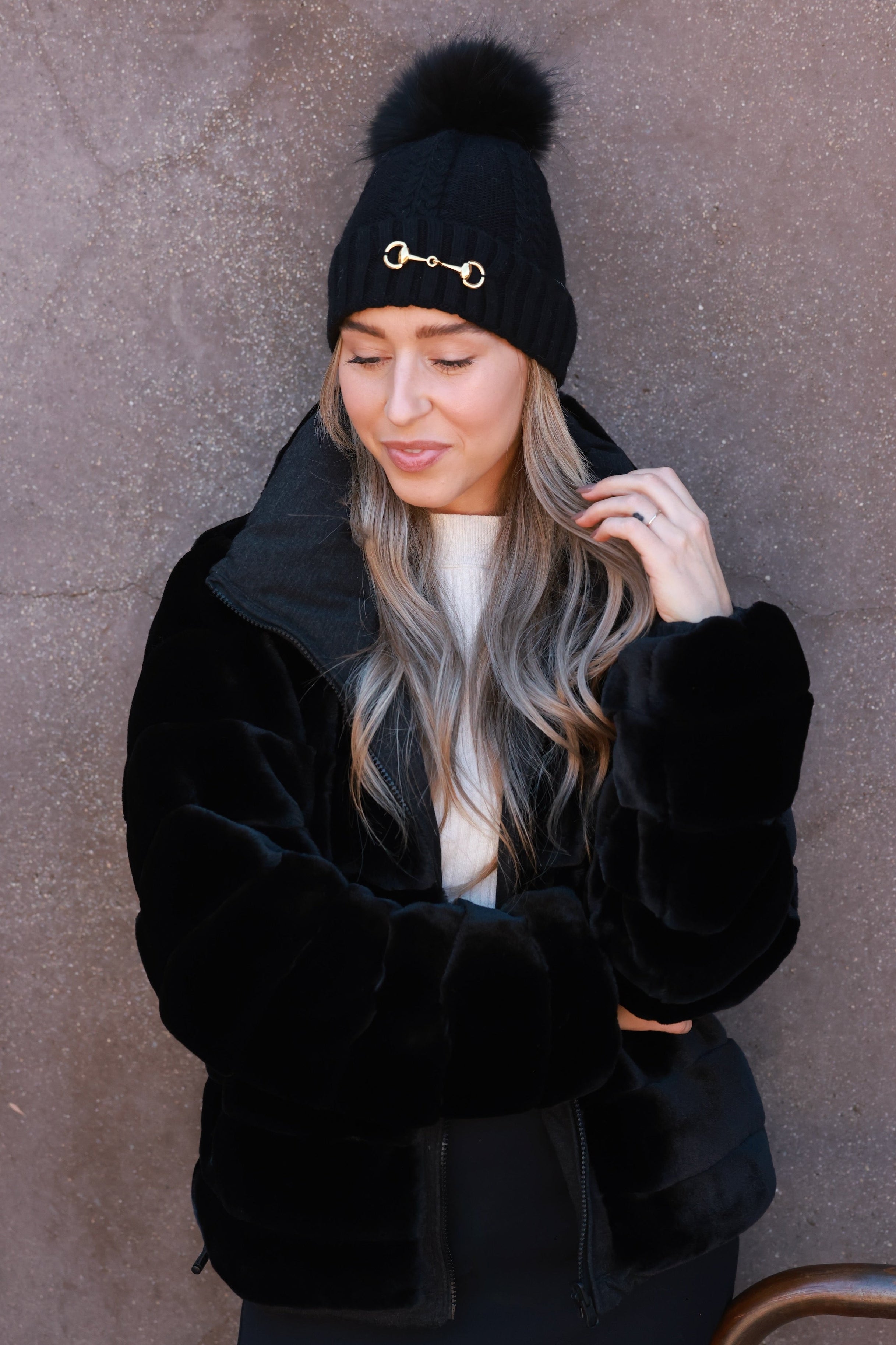 Black Evie Beanie featuring soft, cozy medium-knit cable design with polished gold bit accent and removable faux fur pom. A stylish equestrian winter accessory that adds a chic touch to cold days.
