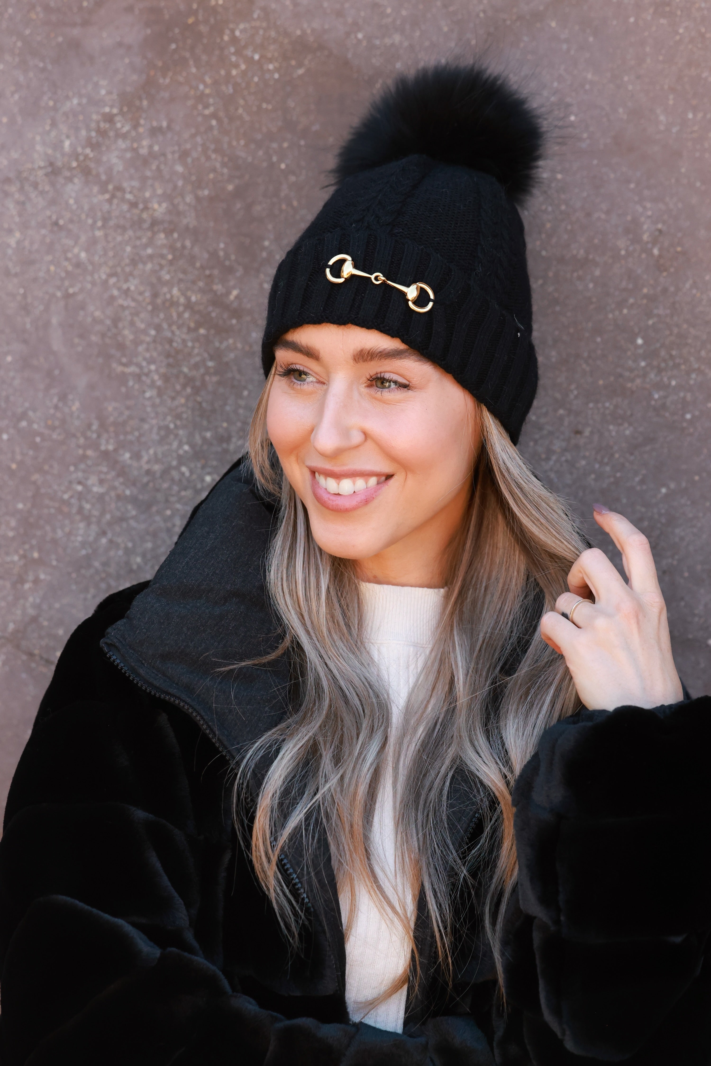 Black Evie Beanie featuring soft, cozy medium-knit cable design with polished gold bit accent and removable faux fur pom. A stylish equestrian winter accessory that adds a chic touch to cold days.