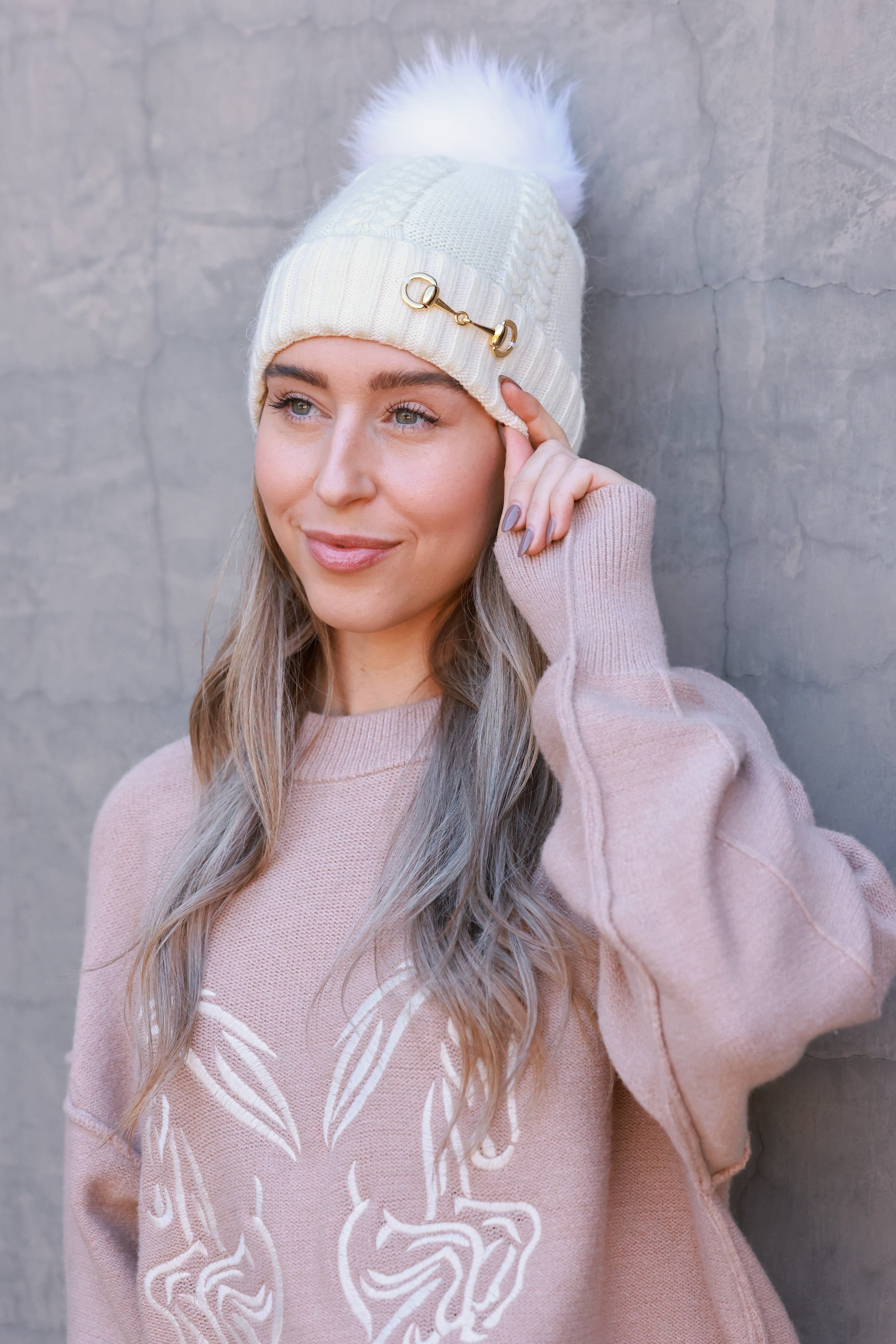 Ivory Evie Beanie featuring a soft, cozy medium-knit cable design with a polished gold bit accent and removable faux fur pom. A timeless equestrian winter accessory that blends warmth with chic style.