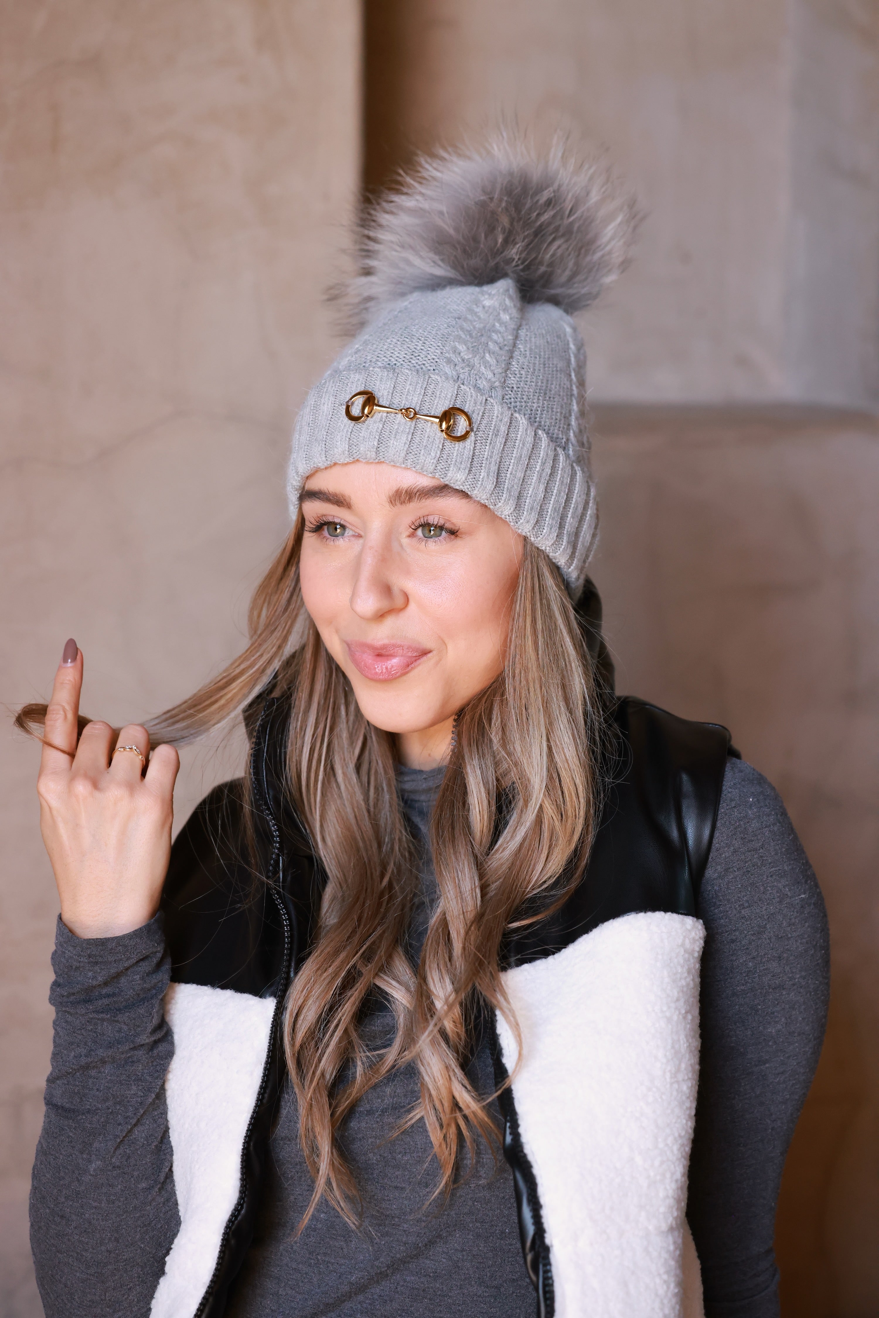 Light grey Evie Beanie featuring soft, cozy medium-knit cable design with polished gold bit accent and removable faux fur pom. A chic equestrian winter accessory adding warmth and style to cool days.