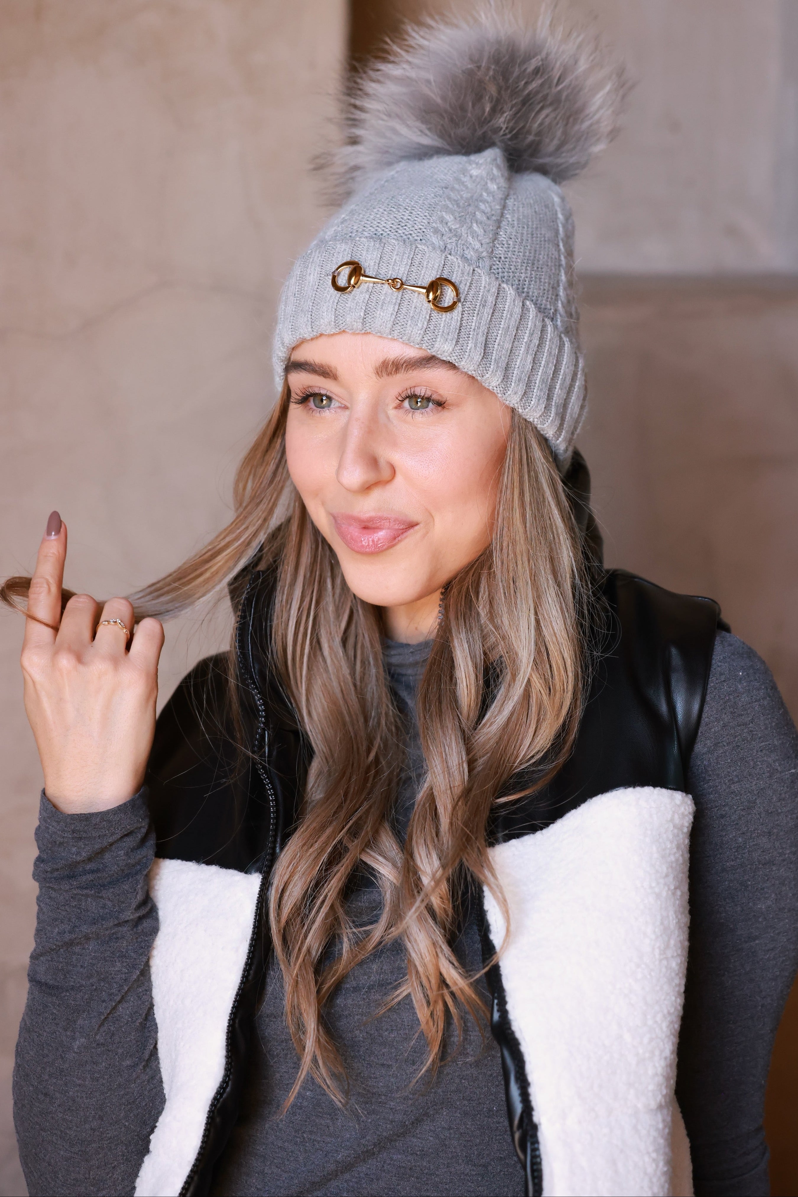 Light grey Evie Beanie featuring soft, cozy medium-knit cable design with polished gold bit accent and removable faux fur pom. A chic equestrian winter accessory adding warmth and style to cool days.