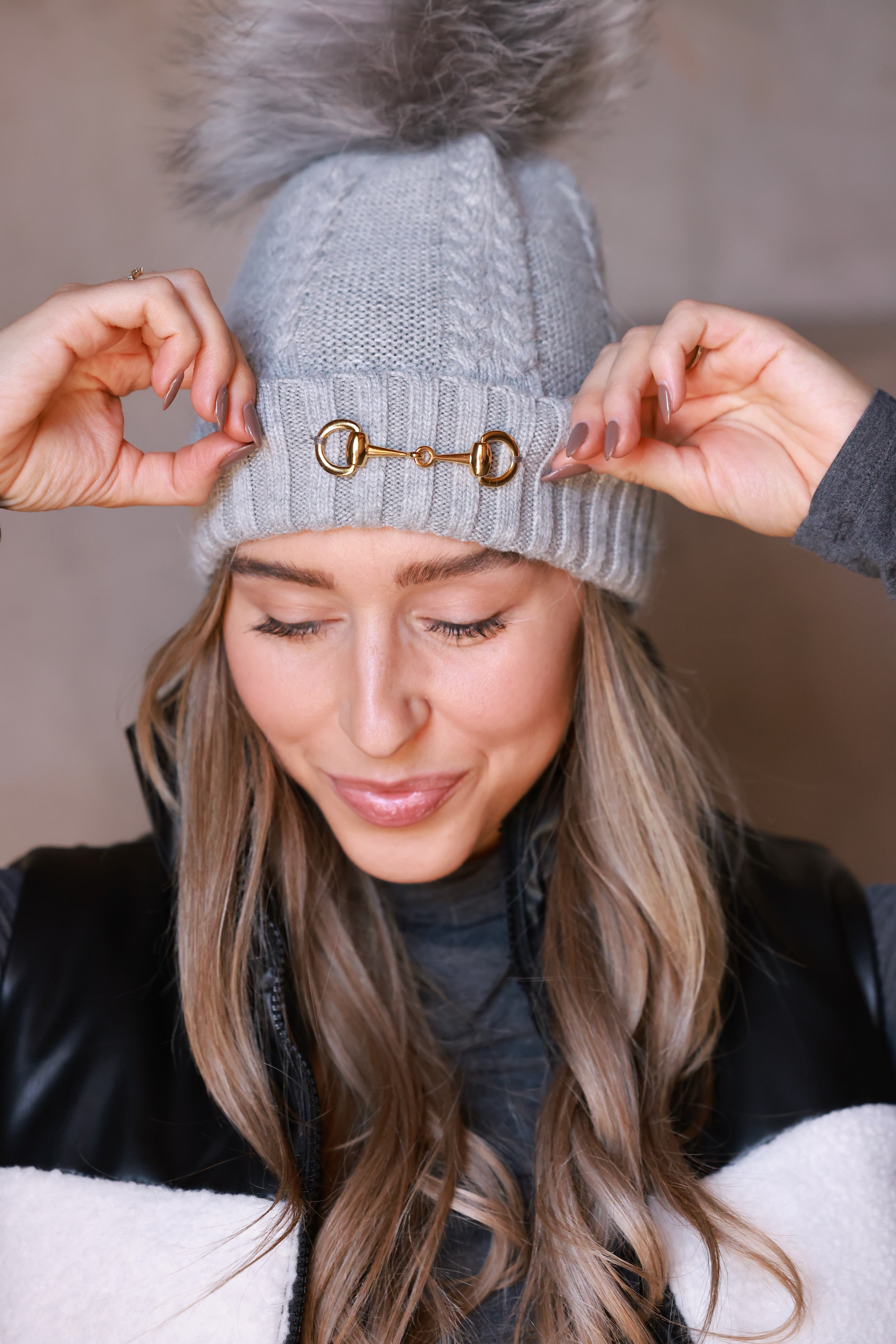 Light grey Evie Beanie featuring soft, cozy medium-knit cable design with polished gold bit accent and removable faux fur pom. A chic equestrian winter accessory adding warmth and style to cool days.