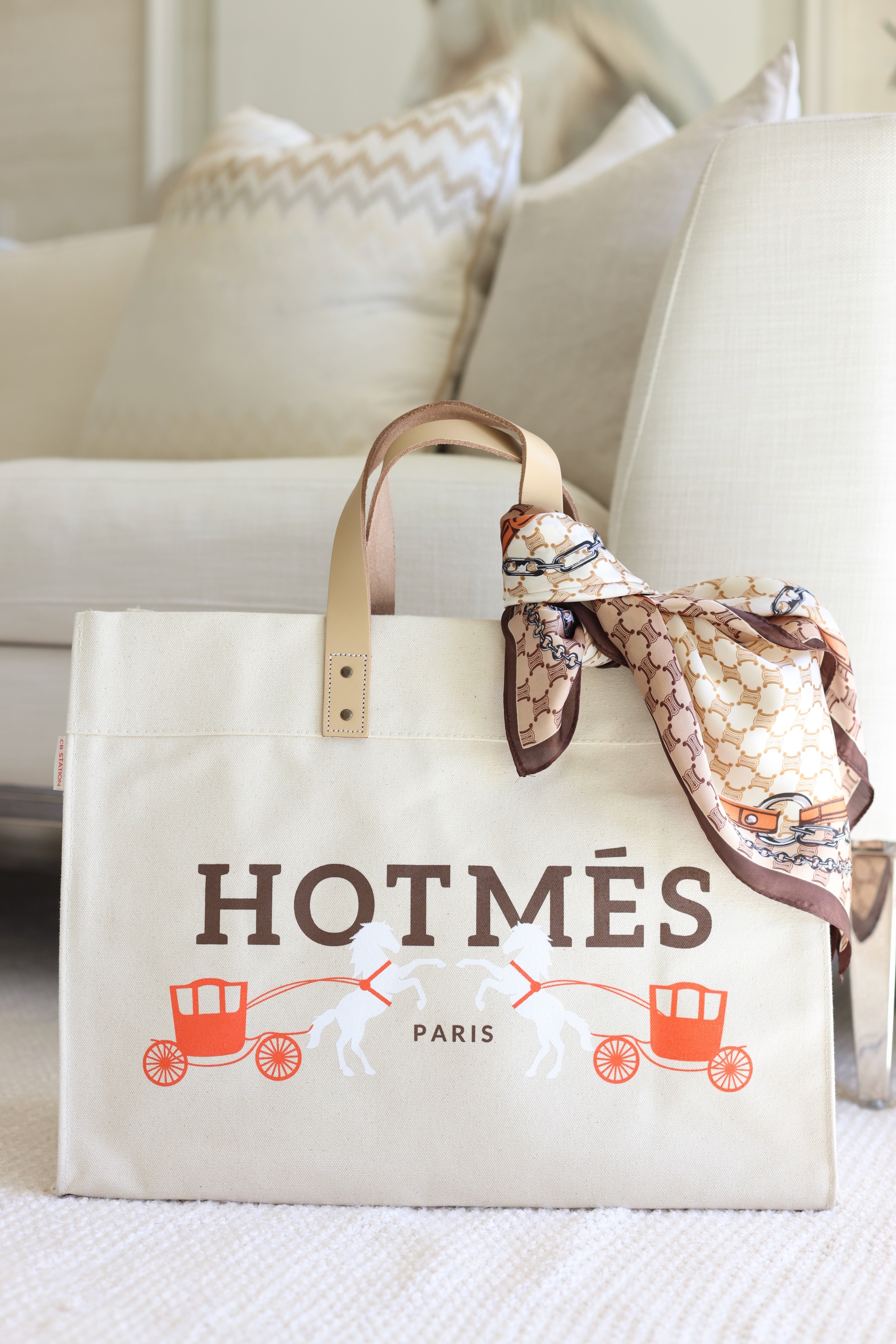 CB Station - A beige tote bag labeled "HOTMÉS" with an orange carriage design and white horses sits on a light carpet. A patterned scarf is tied to its handle, creating an elegant look.