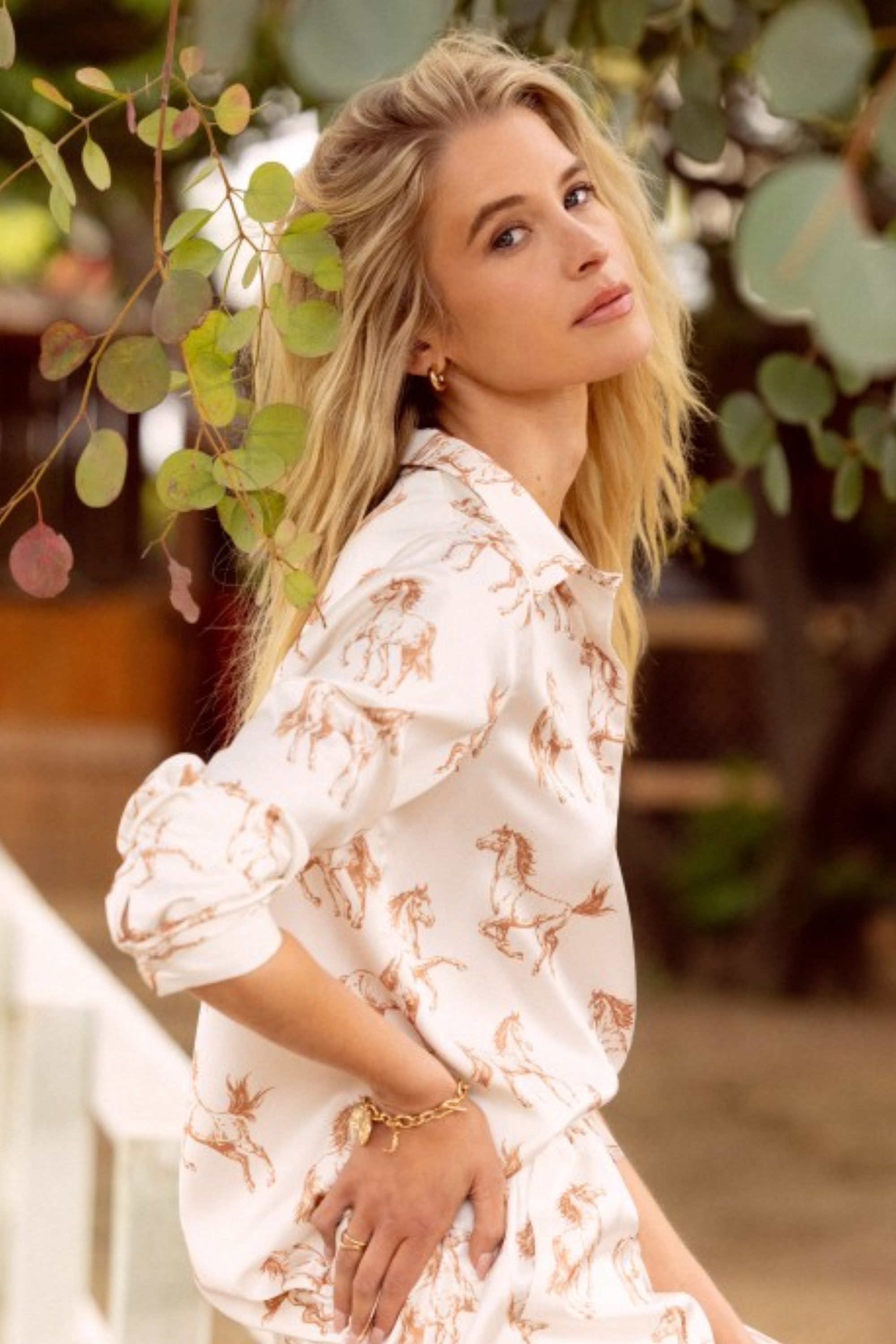 Woman wearing a light, button-down blouse with an all-over horse print, featuring a tie-collar detail and cuffed sleeves. Feminine western riding–themed equestrian top perfect for horse lovers, cowboy-inspired outfits, and romantic horse girl apparel.