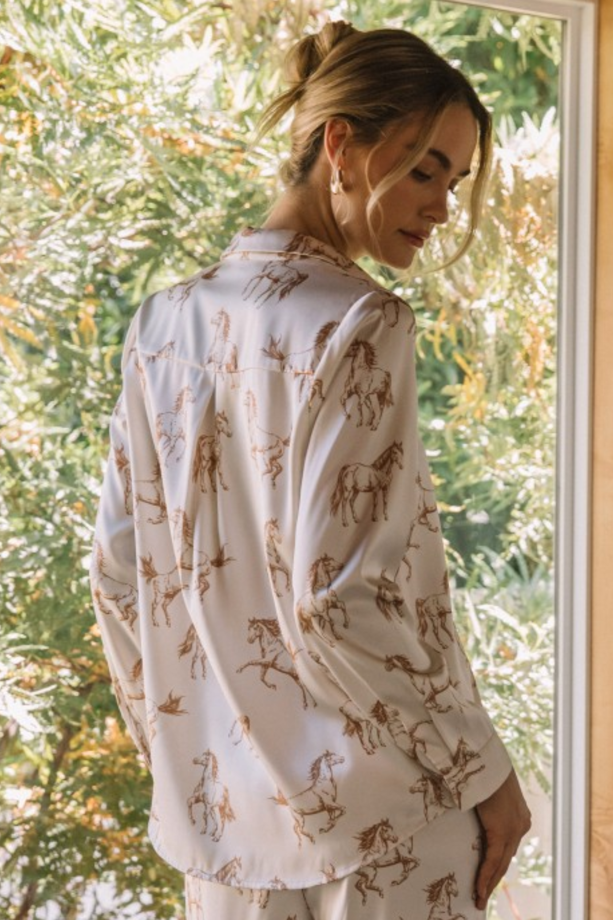 Woman wearing a light, button-down blouse with an all-over horse print, featuring a tie-collar detail and cuffed sleeves. Feminine western riding–themed equestrian top perfect for horse lovers, cowboy-inspired outfits, and romantic horse girl apparel.