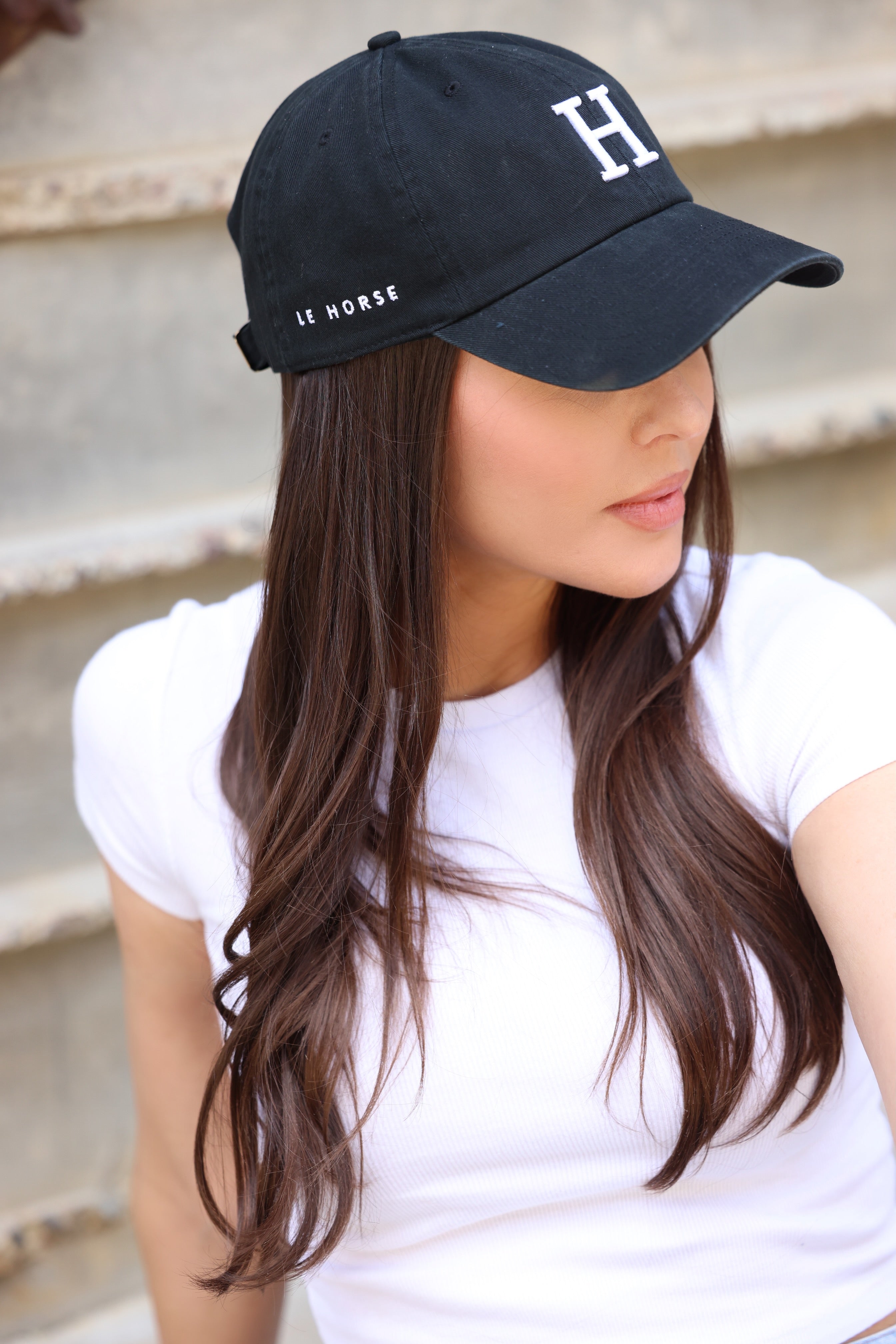 Woman in a white t-shirt and black cap with "H" and "Le Horse," long brown hair visible, sits on steps, looking to the side with a neutral expression.
