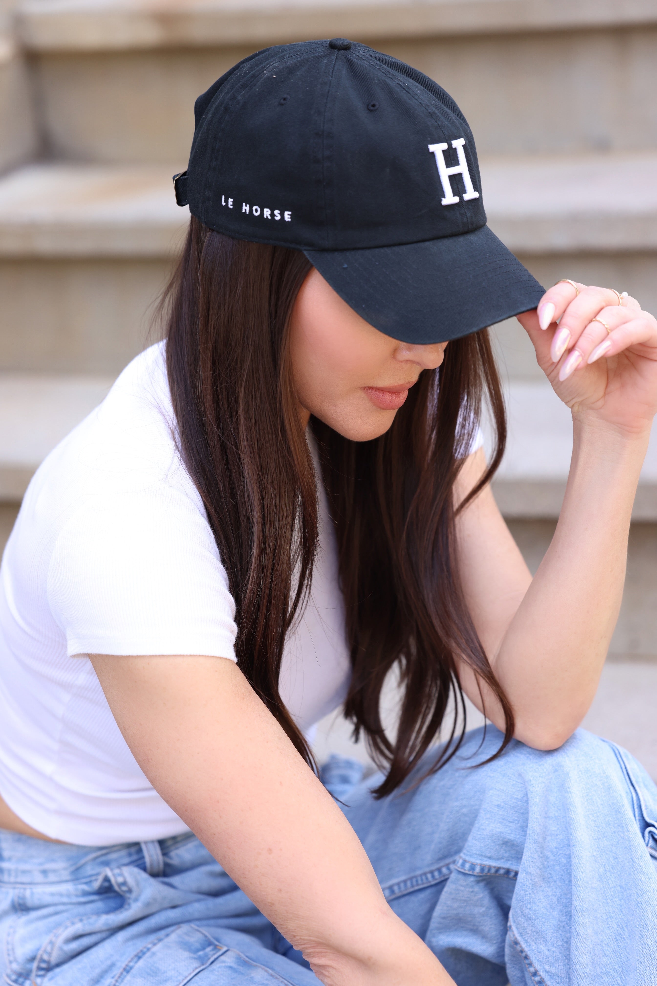 A woman sits on steps, wearing a black cap with "H" and "LE HORSE" text, white shirt, and jeans. She gently holds her cap's brim. Casual and relaxed tone.
