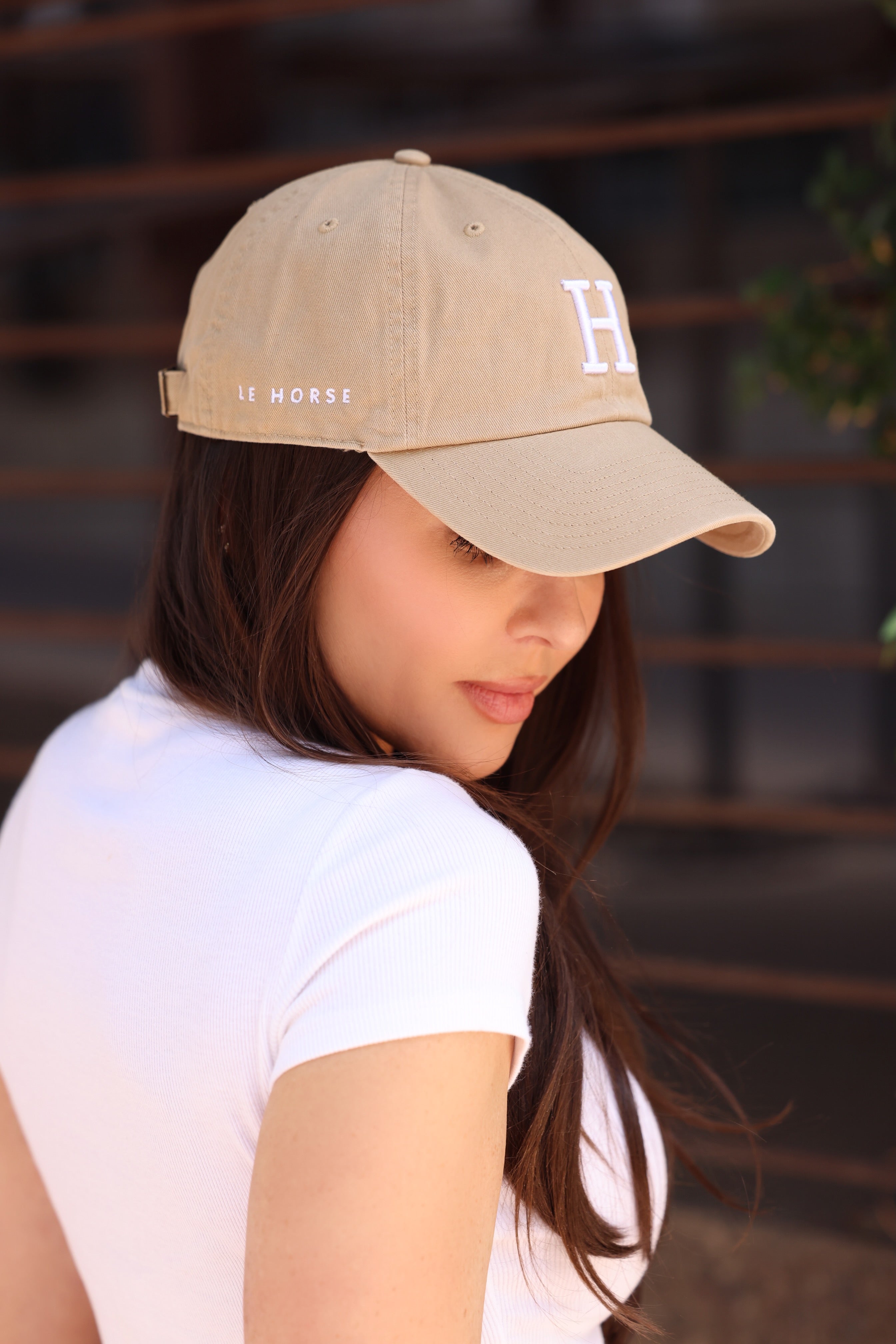 A woman in a white shirt and beige baseball cap with "H" and "Le Horse" embroidery looks downward, set against a blurred background, conveying a serene mood.