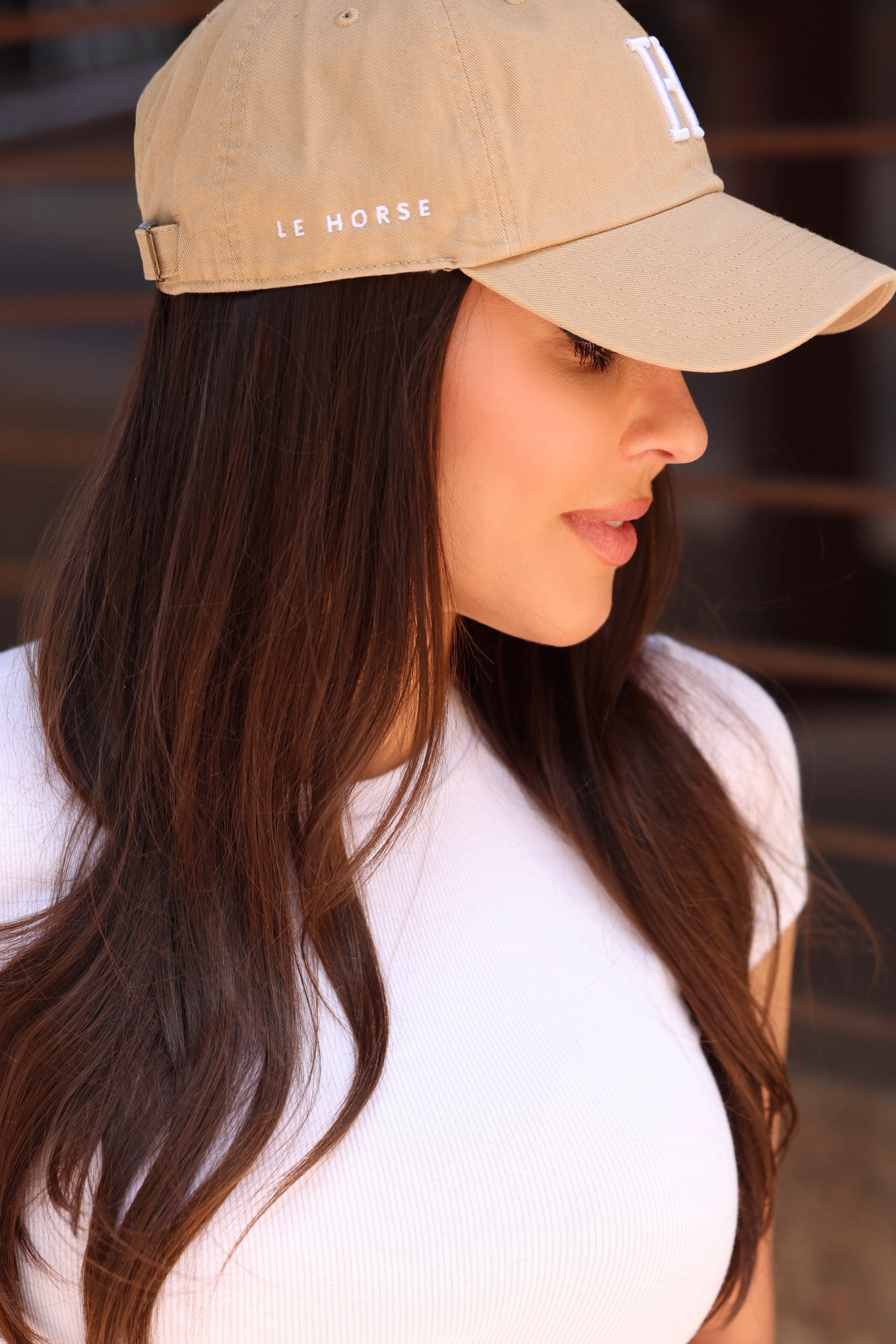 A woman wearing a beige cap with "Le Horse" text and a white shirt, looking down thoughtfully, with long brown hair cascading over her shoulders.
