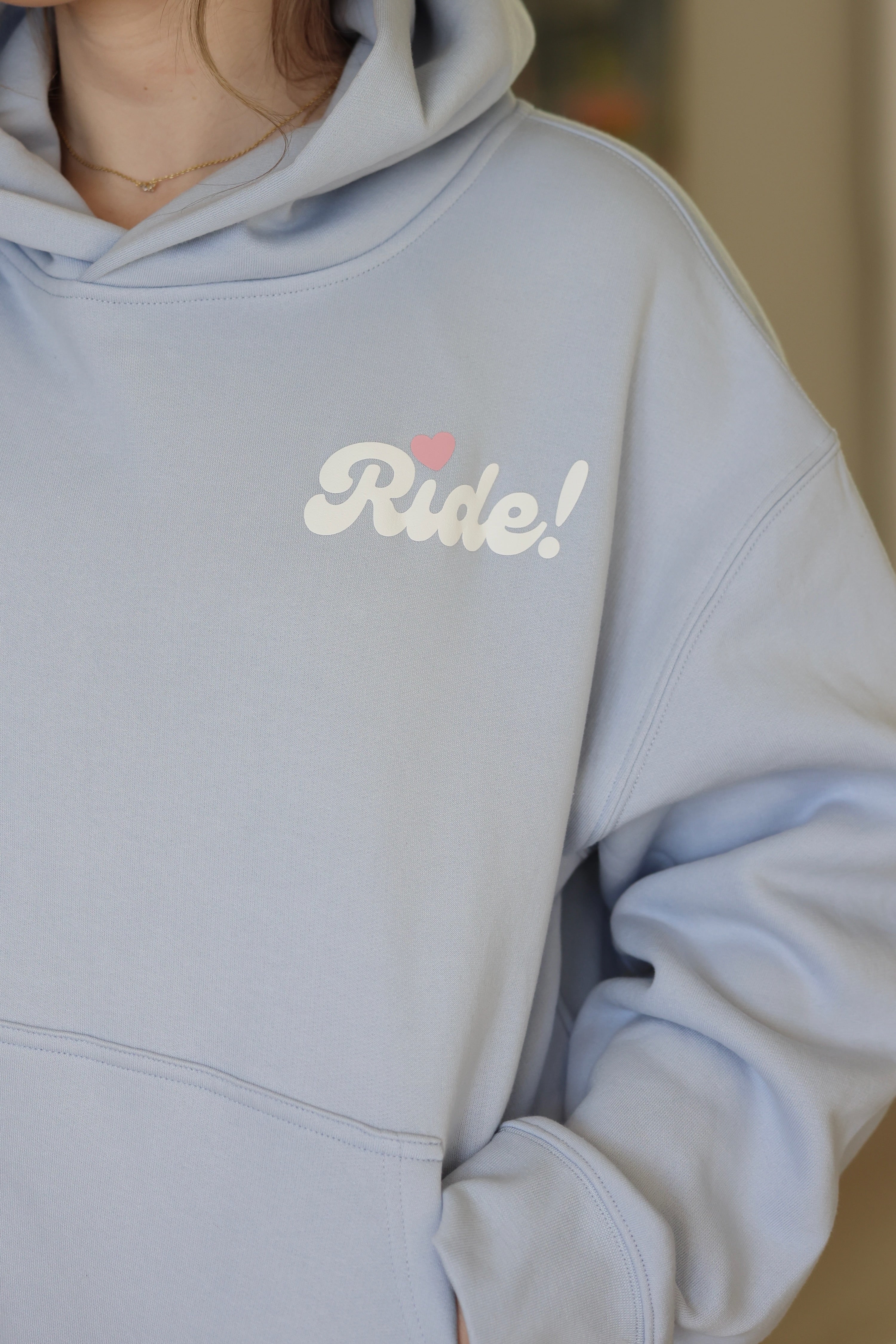 A person wearing a light blue hoodie with "Ride!" and a small pink heart printed on it, conveying a casual and playful tone.