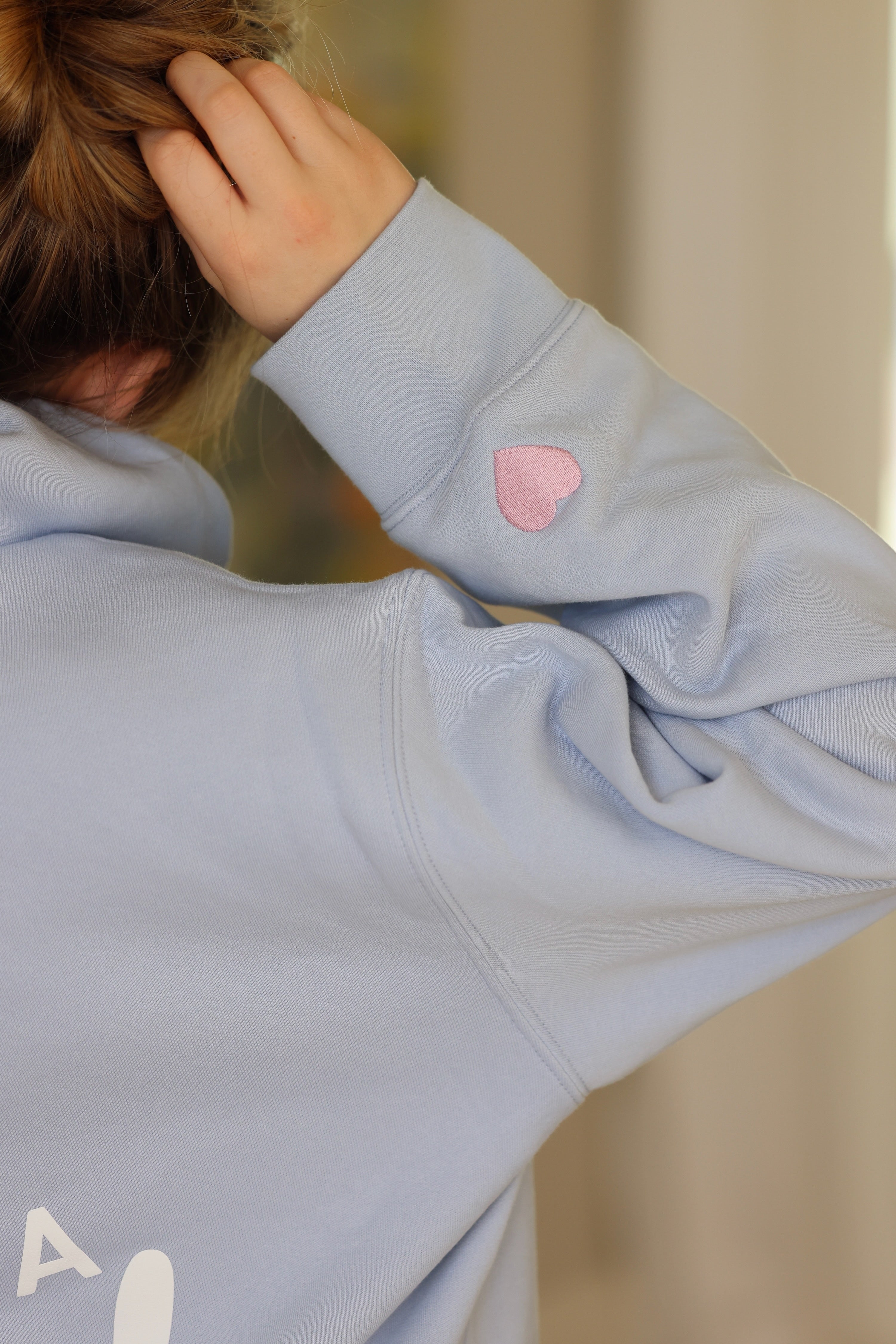 The image shows a person from behind wearing a light blue sweatshirt with a pink heart patch on the sleeve. They are adjusting their hair.