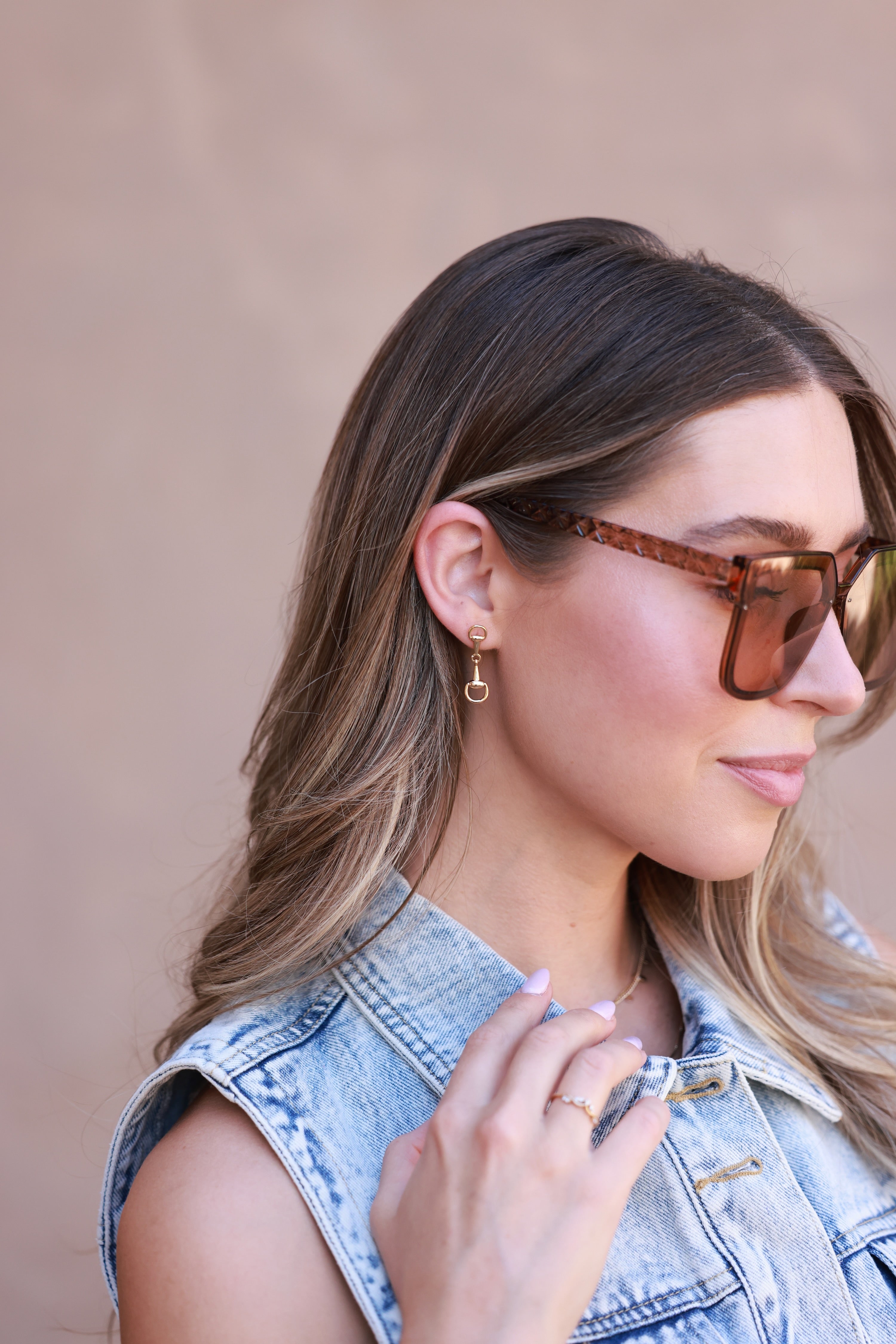 Woman wearing sunglasses and a denim vest against a beige background - horse bit earrings, equestrian inspired jewelry, horse bit jewelry, equestrian accessories, dainty equestrian jewelry, snaffle bit earrings, horse girl jewelry, horse girl accessory