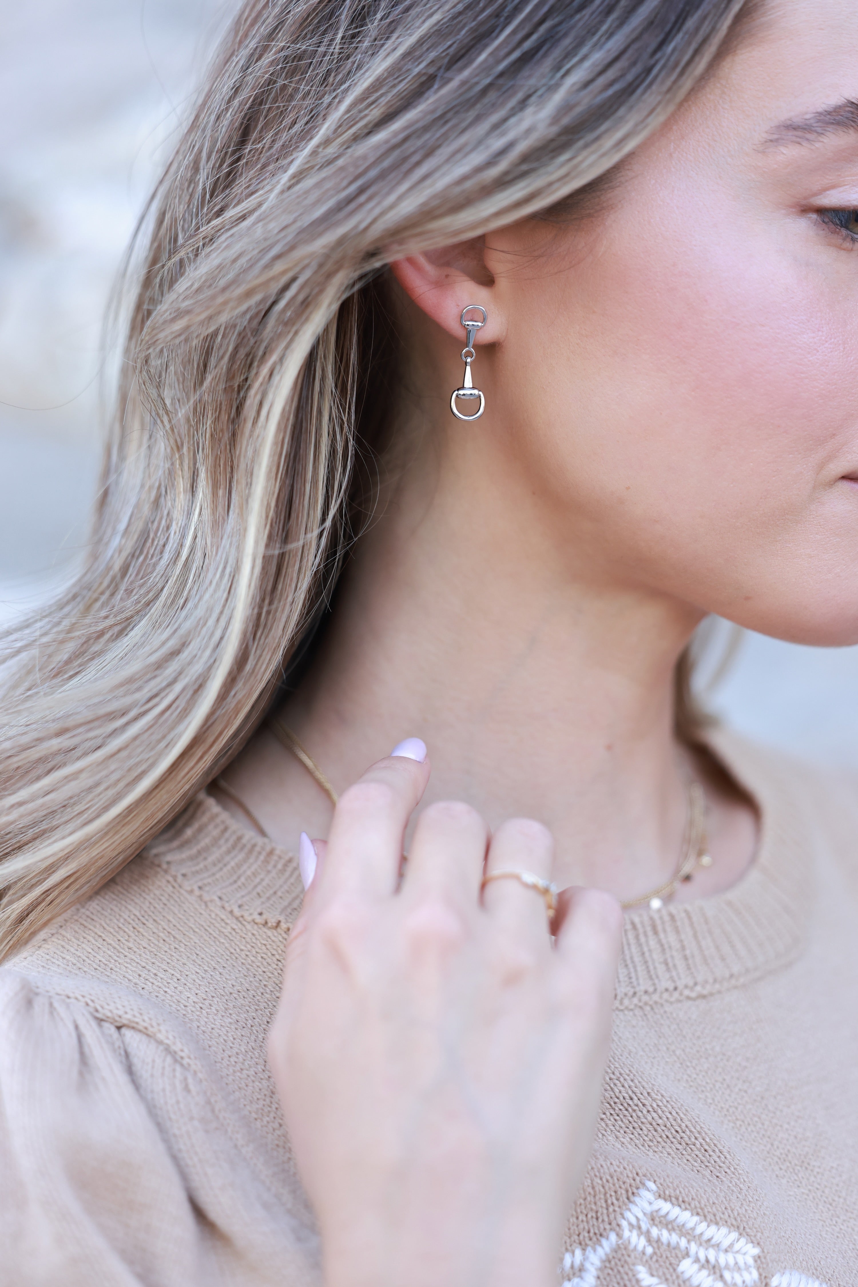 Close-up of a woman wearing silver earrings and a ring, with a neutral background. - horse bit earrings, equestrian inspired jewelry, horse bit jewelry, equestrian accessories, dainty equestrian jewelry, snaffle bit earrings, horse girl jewelry, horse girl accessory