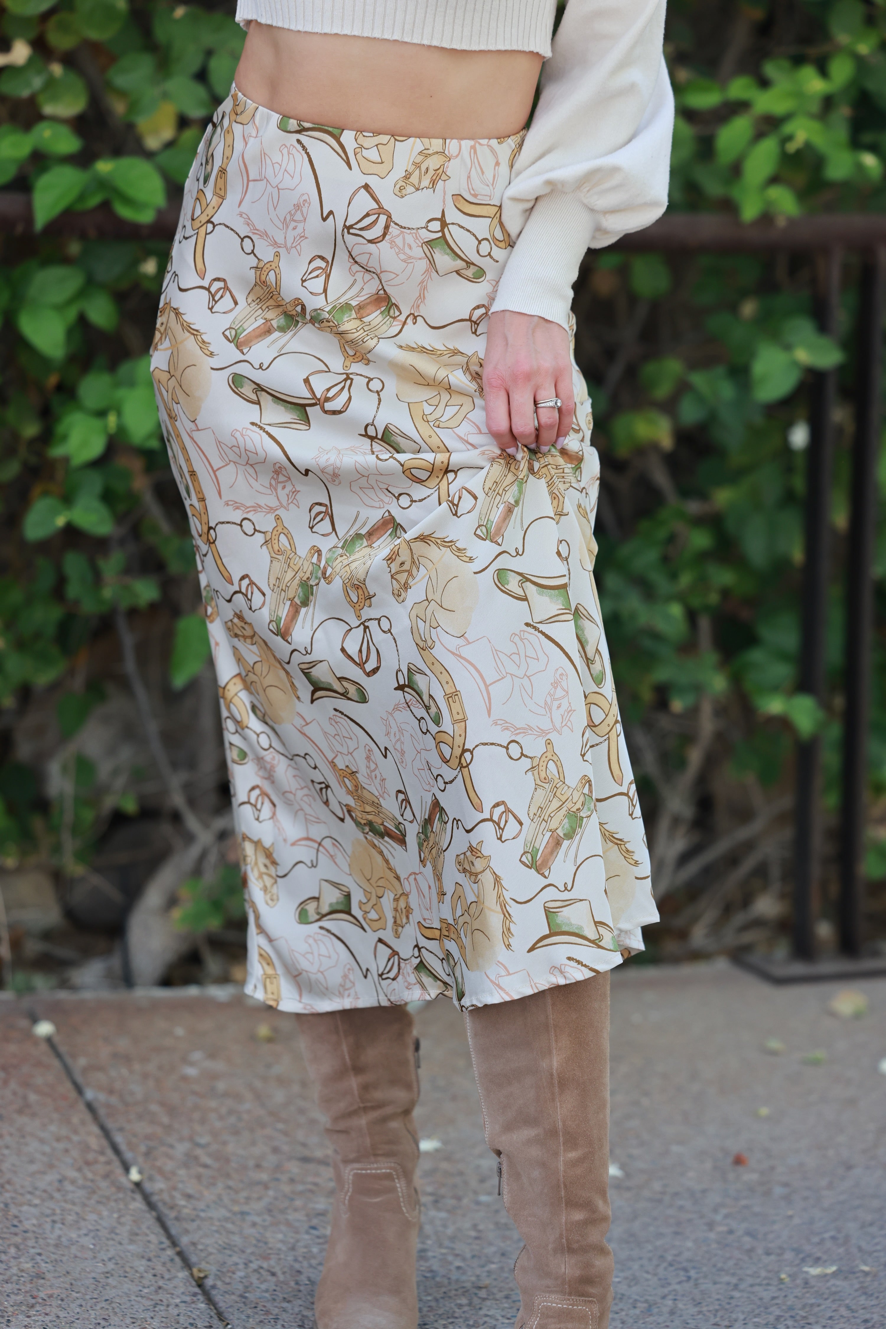 Person wearing a patterned skirt with a blurred green background - Western satin midi skirt, &merci exclusive, Cowgirl print slip skirt, Equestrian-inspired fashion, Fall rodeo outfit