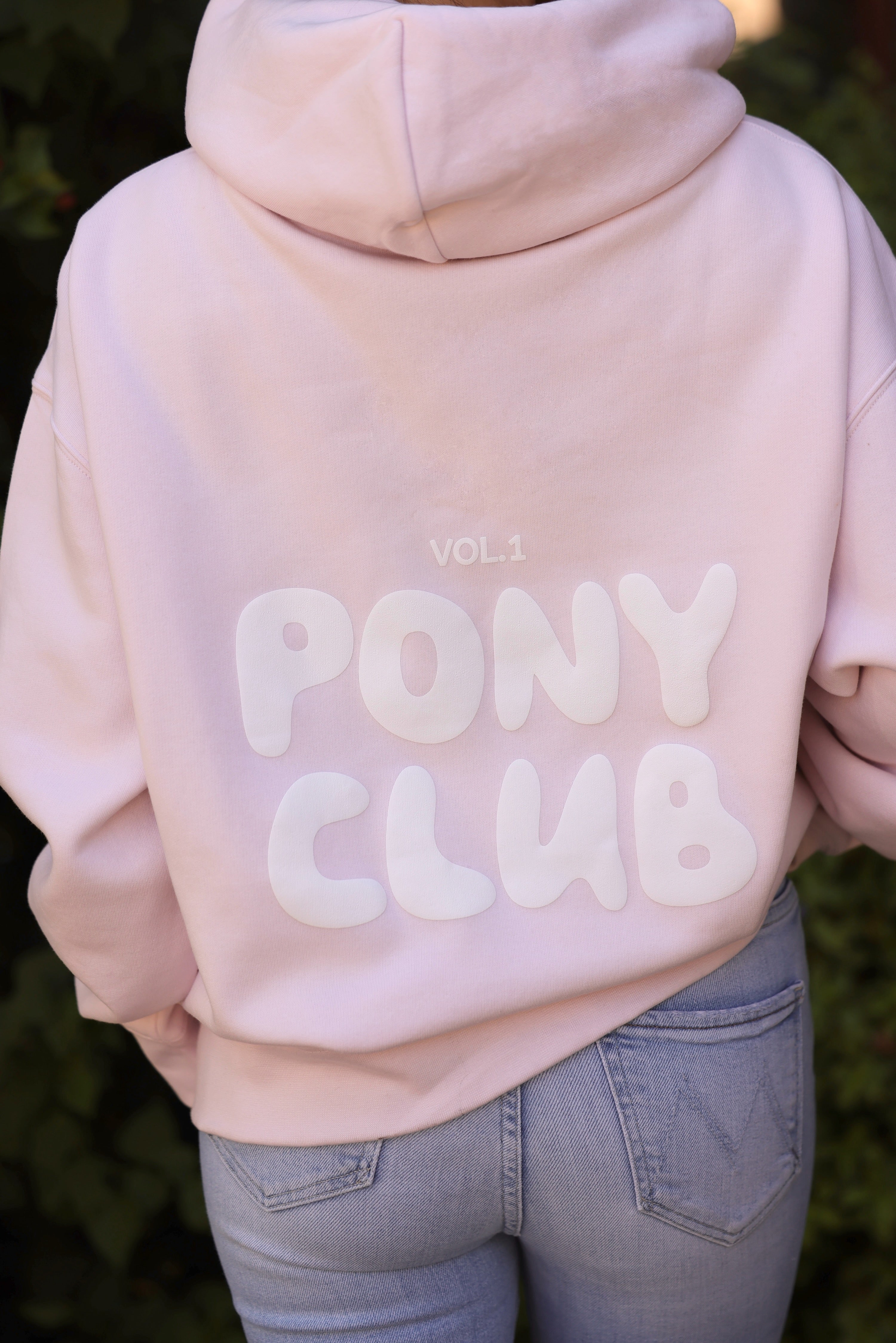 Rear view of a person wearing a pastel petal pink hoodie with "Vol 1 PONY CLUB" printed in bold white letters. The setting is casual and relaxed.