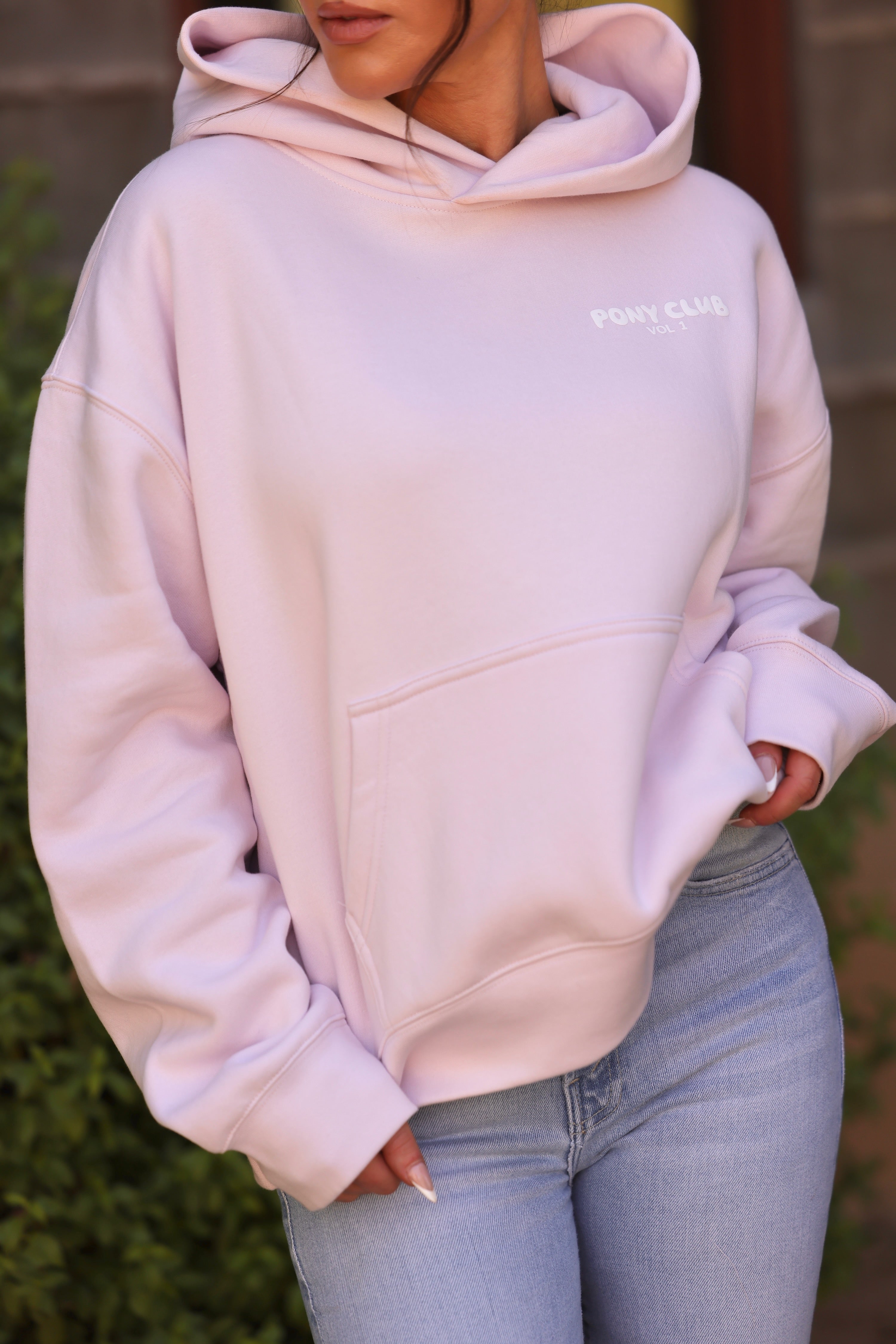 Close-up of a person wearing a light petal pink hoodie with "PONY CLUB VOL. 1" in white text. The fabric appears soft, conveying a cozy feel.