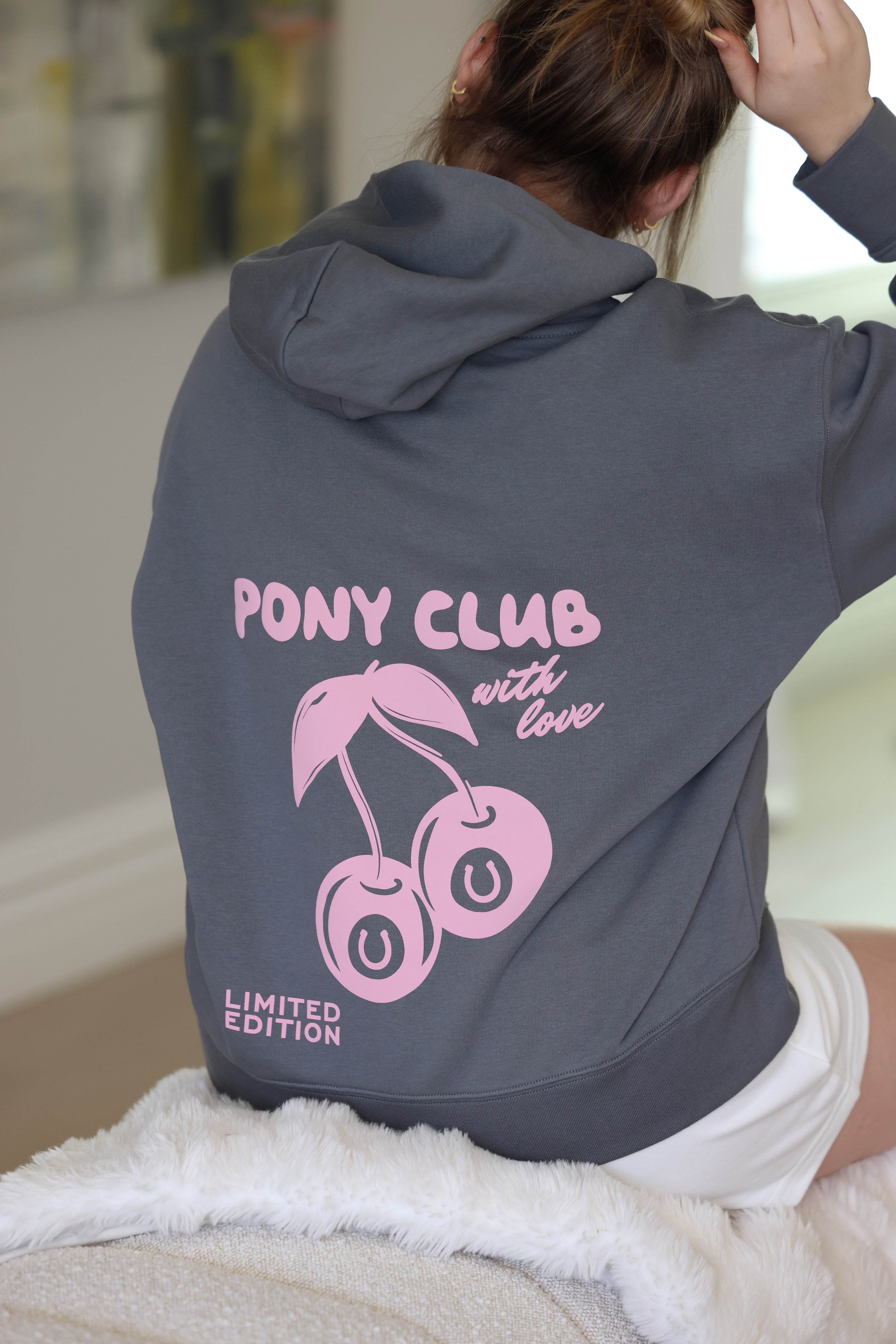 A person seated with their back to the camera, wearing a shadow gray hoodie featuring pink cherries and "PONY CLUB with love" text in a cozy, casual setting.