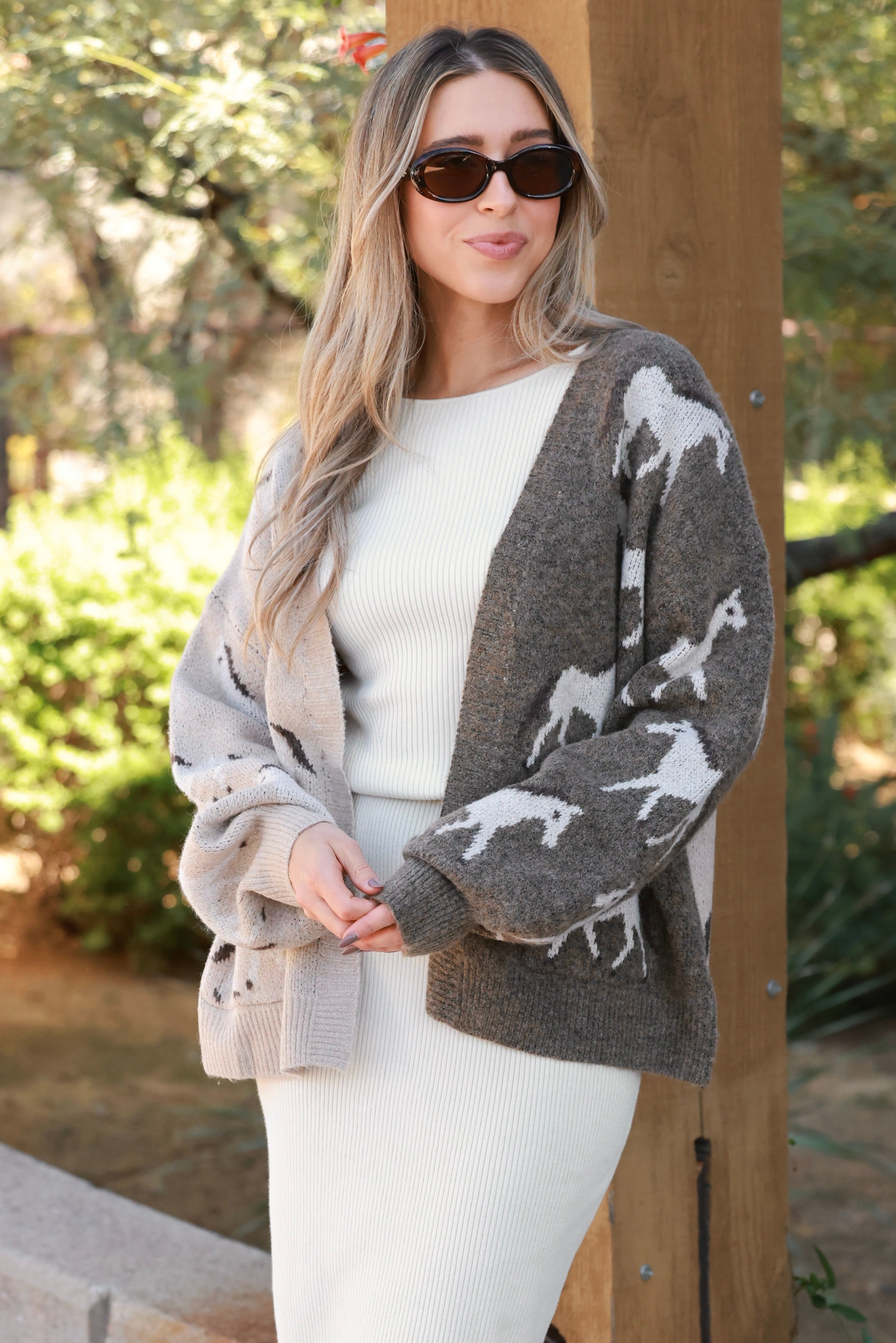 Woman wearing an asymmetric open-front cardigan in brown and beige with white horse patterns. This cozy equestrian cardigan blends rustic western style with casual horse girl apparel, soft, stretchy knit perfect for everyday wear.
