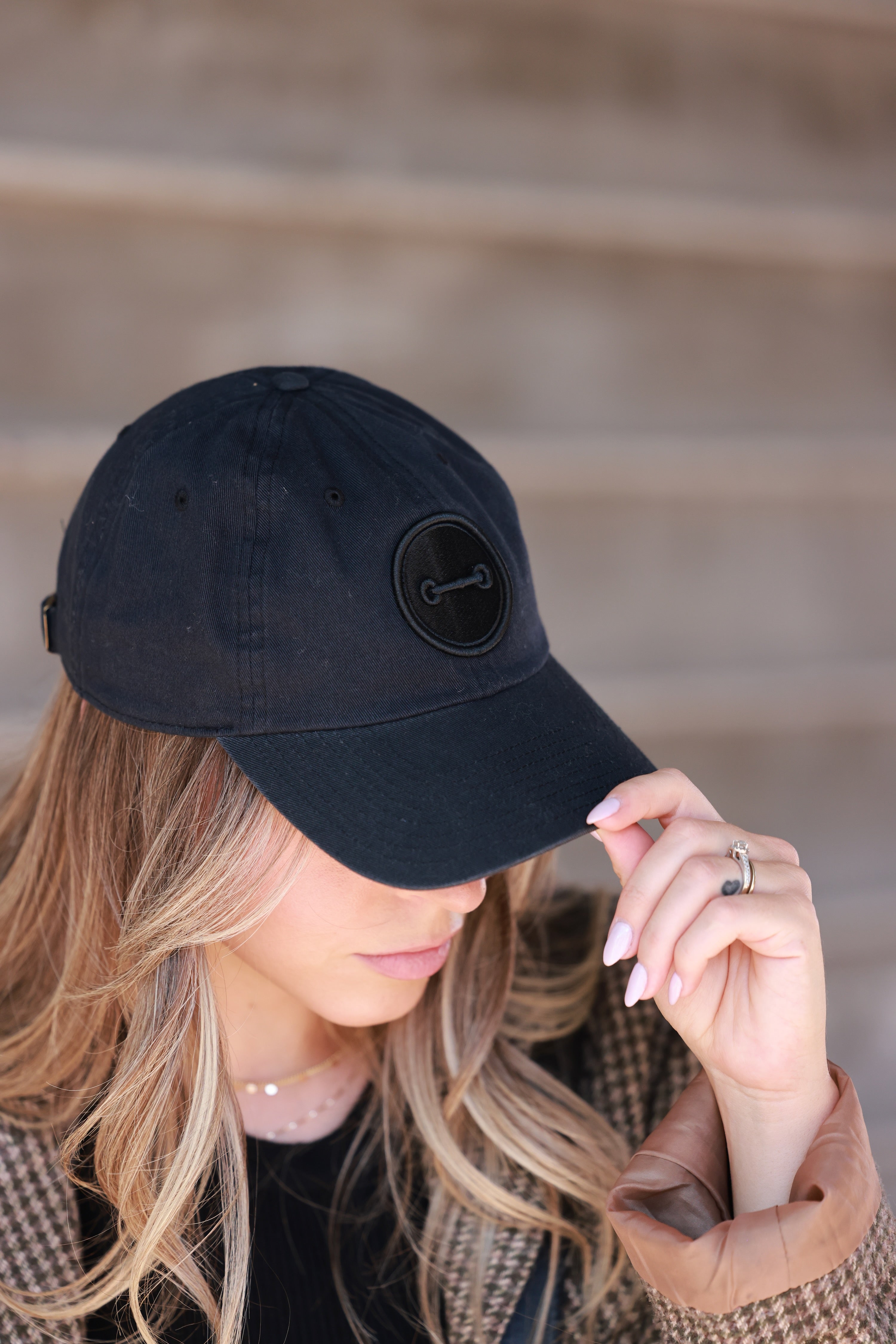 Woman wearing a black cap with a logo, adjusting it against a blurred background - horse bit hat, equestrian headwear, horse girl accessories, casual riding gear, horse riding accessories, baseball cap