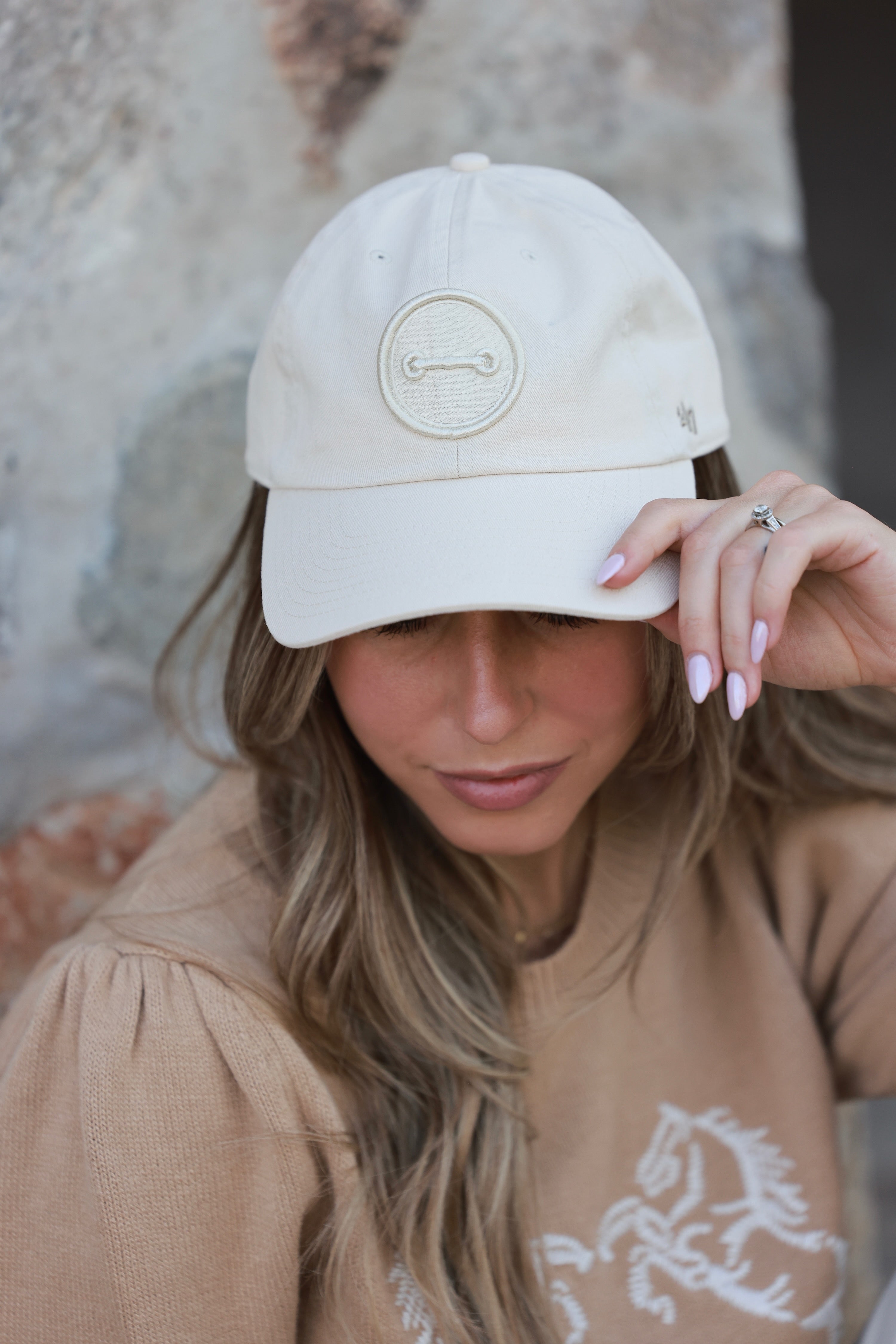 Woman wearing a beige cap and sweater with a horse design, sitting against a stone wall. - horse bit hat, equestrian headwear, horse girl accessories, casual riding gear, horse riding accessories, baseball cap
