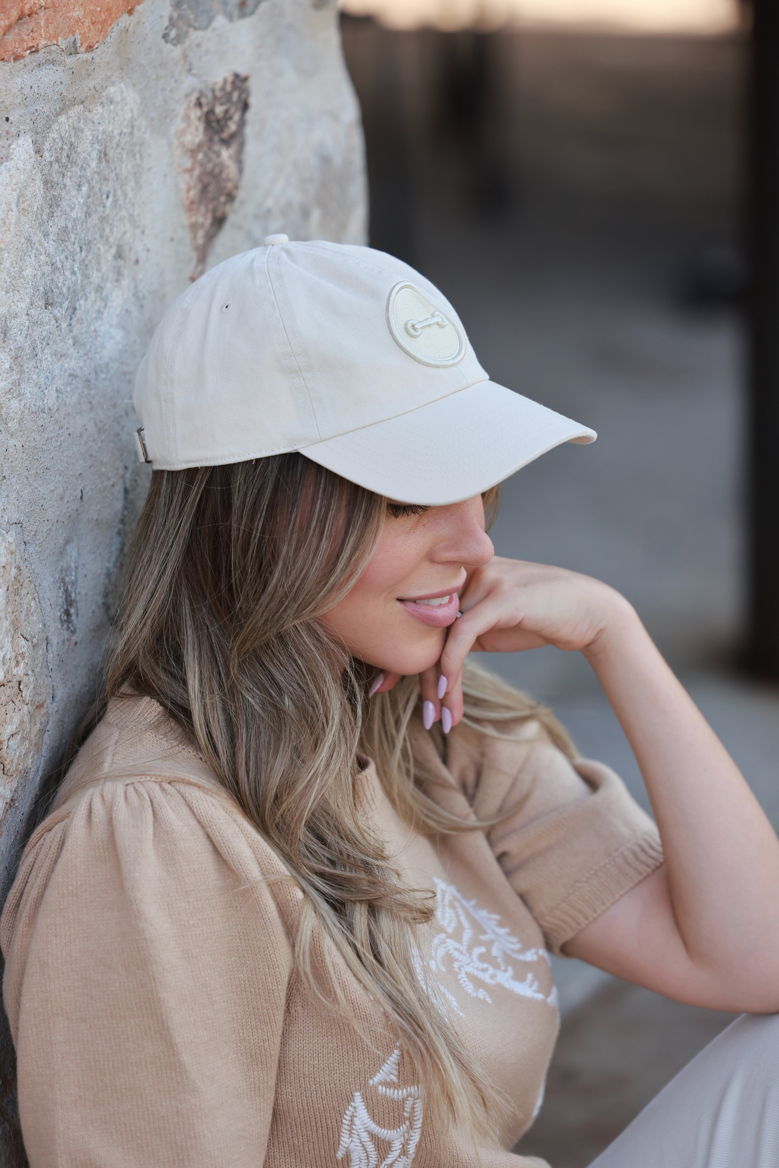 Woman wearing a beige cap and shirt, leaning against a stone wall. - horse bit hat, equestrian headwear, horse girl accessories, casual riding gear, horse riding accessories, baseball cap