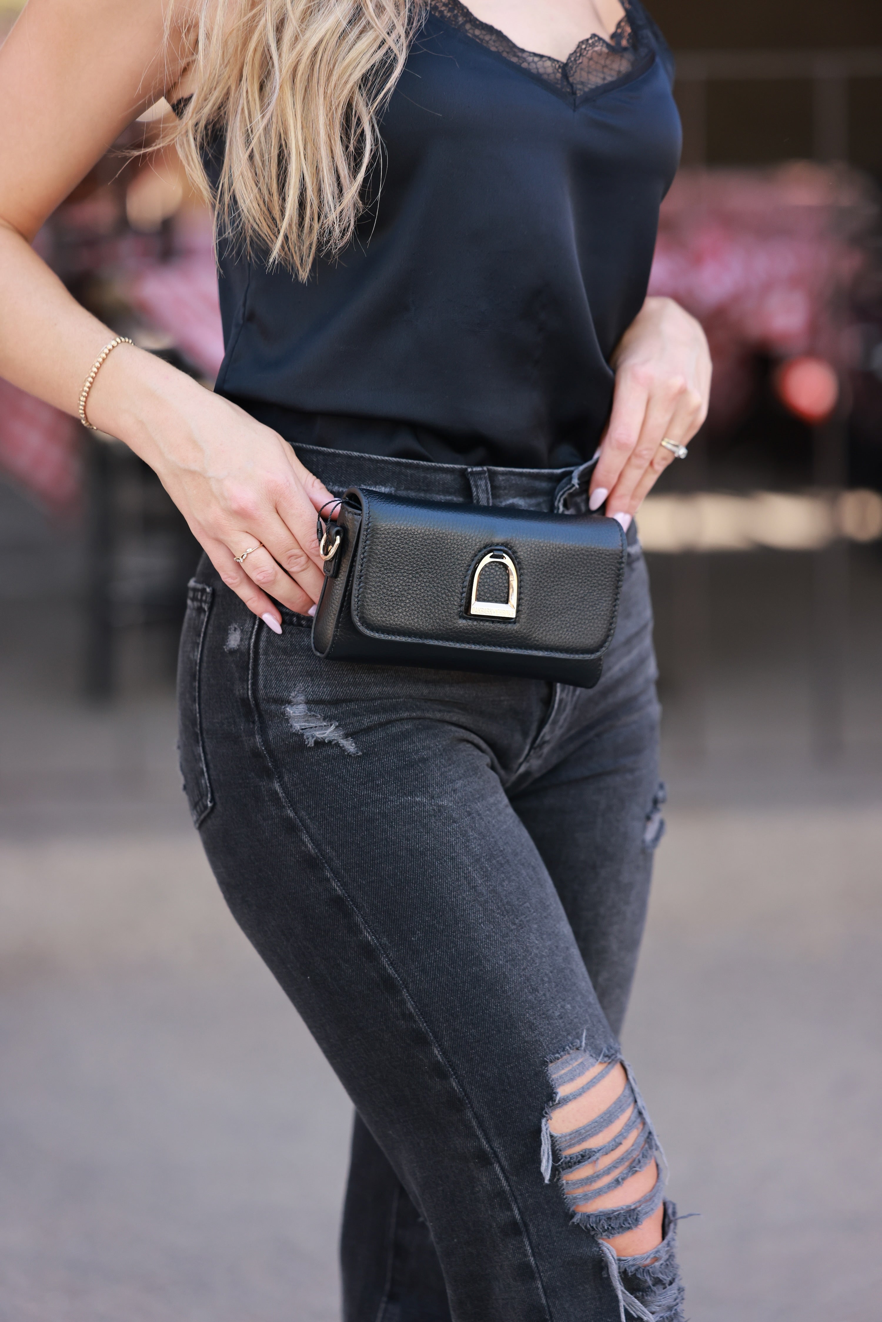 Person wearing a black sleeveless top and ripped jeans, holding a black handbag. - Horse stirrup belt bag, Equestrian-inspired belt bag;stirrup detail belt bag