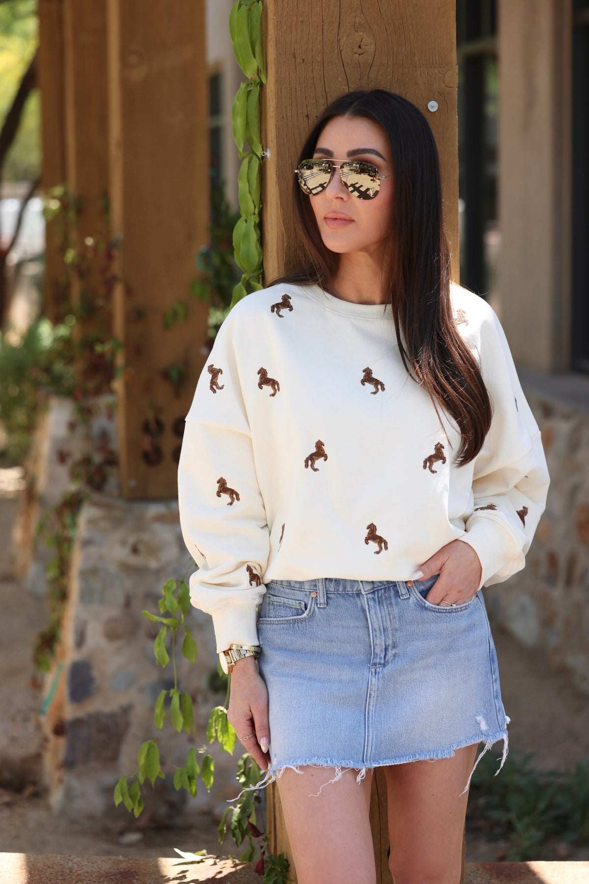 Woman wearing reflective sunglasses, a white sweater with horse patterns, and a denim skirt, standing by a wooden pillar with climbing vines. Casual ambiance.