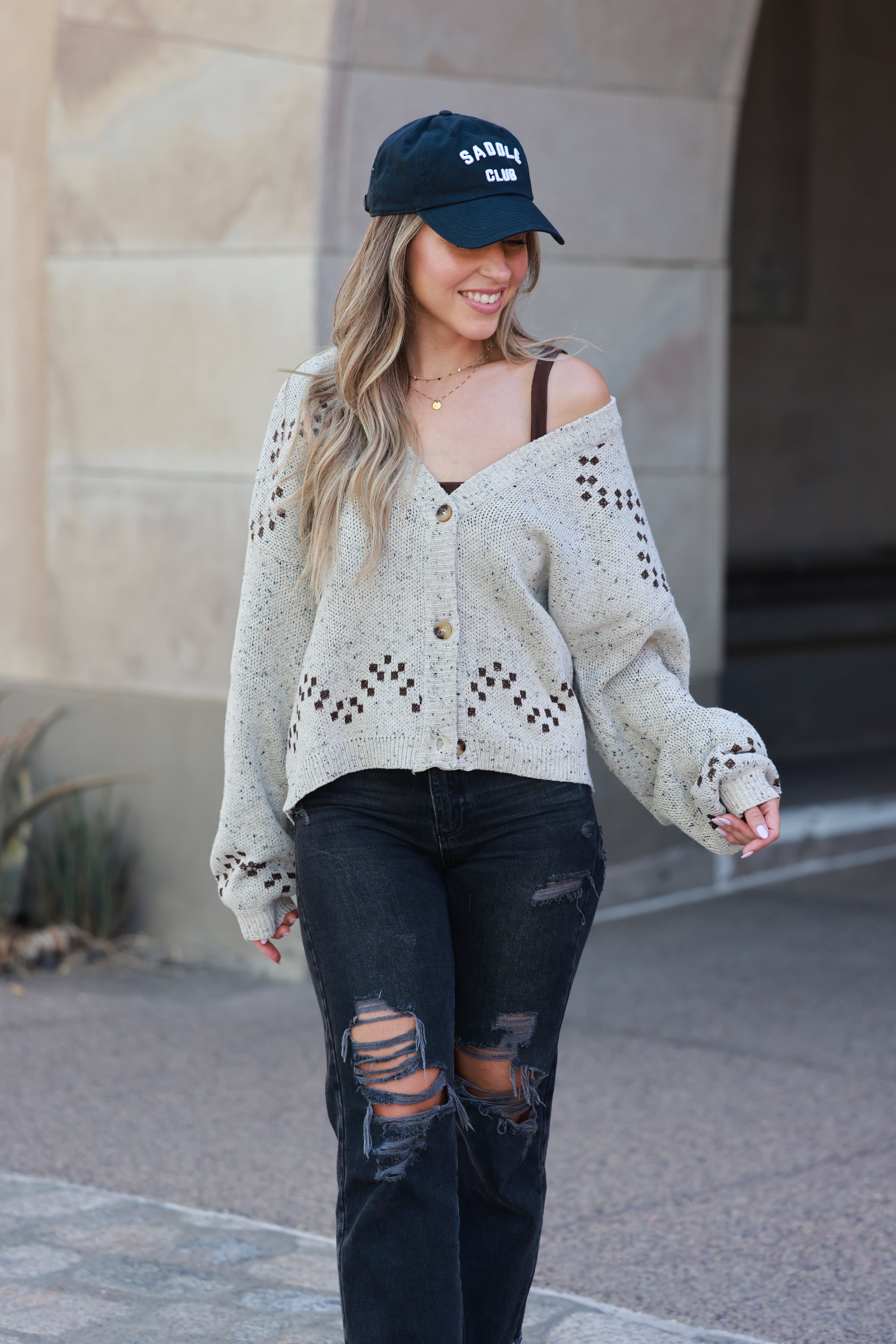 Woman wearing a light-colored cardigan with cut-out details, black distressed jeans, and a dark cap outdoors. Horse graphic cardigan, Western button down sweater, Equestrian fall knit, Oatmeal horse print cardigan, Cowgirl boutique sweater