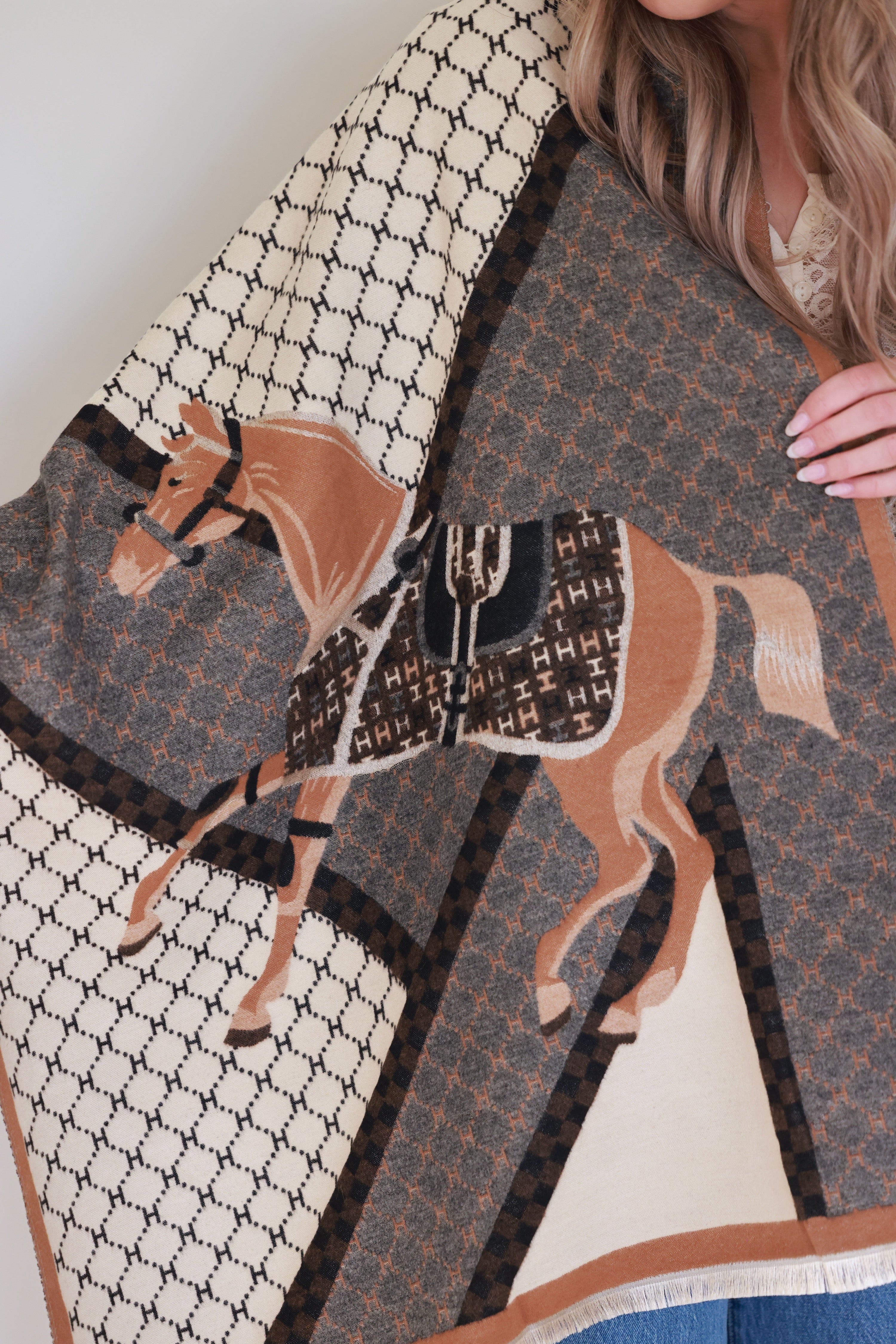 Camel-colored equestrian-inspired scarf with soft cashmere-like texture and subtle horse motif, featuring eyelash fringe and finished hems, styled as a chic wrap for day or evening wear.