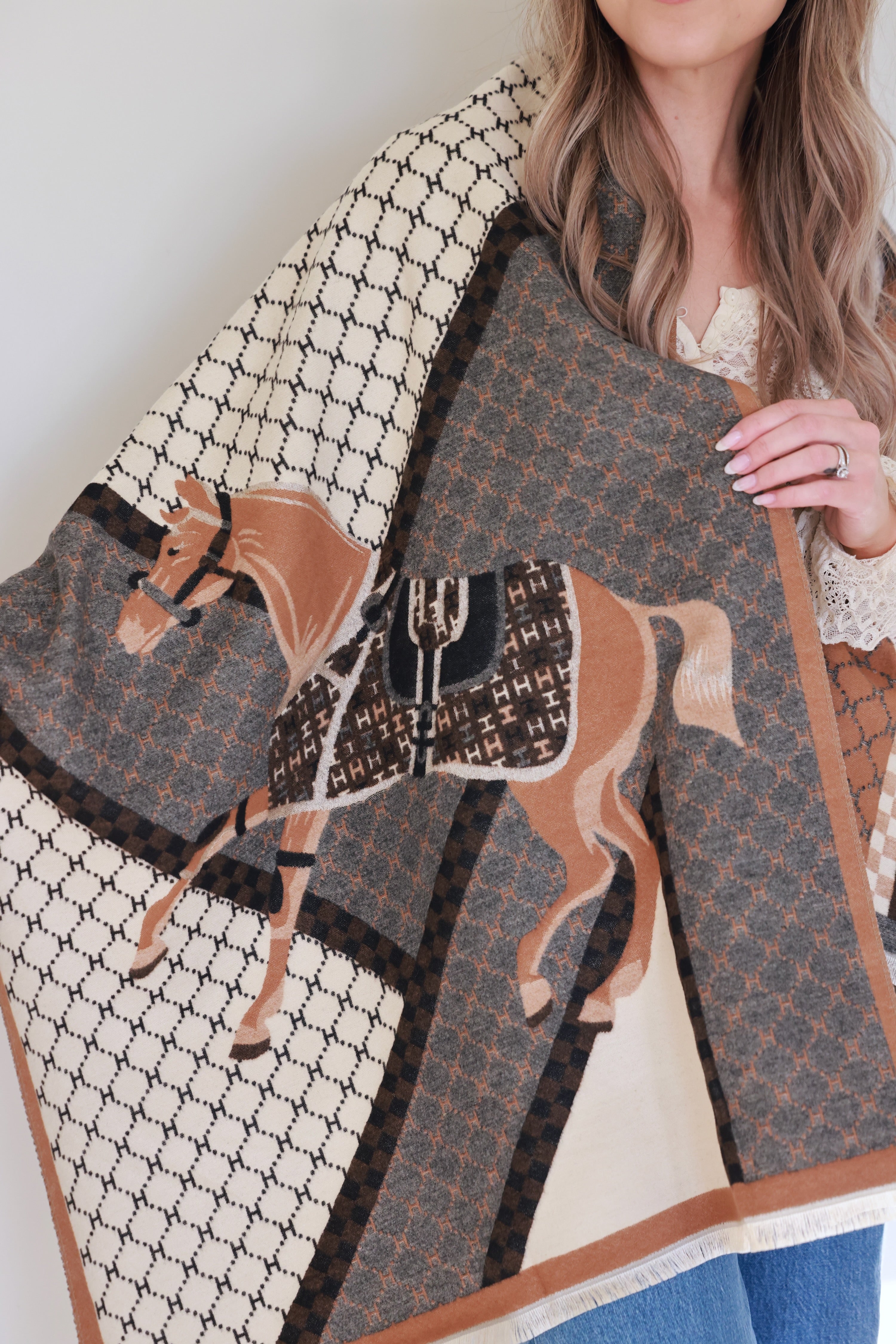 Camel-colored equestrian-inspired scarf with soft cashmere-like texture and subtle horse motif, featuring eyelash fringe and finished hems, styled as a chic wrap for day or evening wear.
