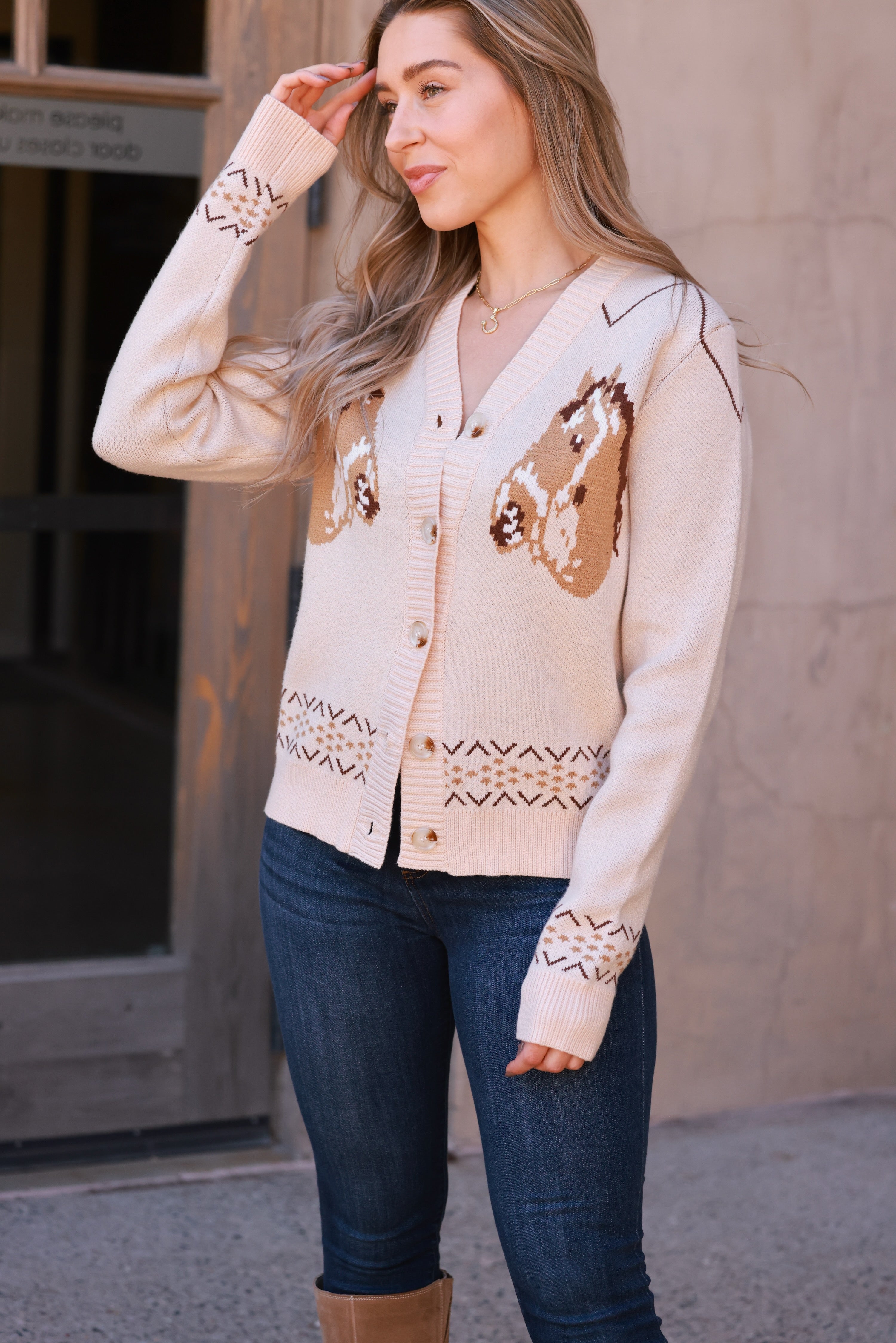A cozy cream-colored knit cardigan with two brown horse graphics and subtle geometric fair isle trim. This long-sleeve, V-neck button-down features a relaxed, slightly cropped fit and soft viscose-poly blend fabric, offering a playful, equestrian-inspired look perfect for fall.