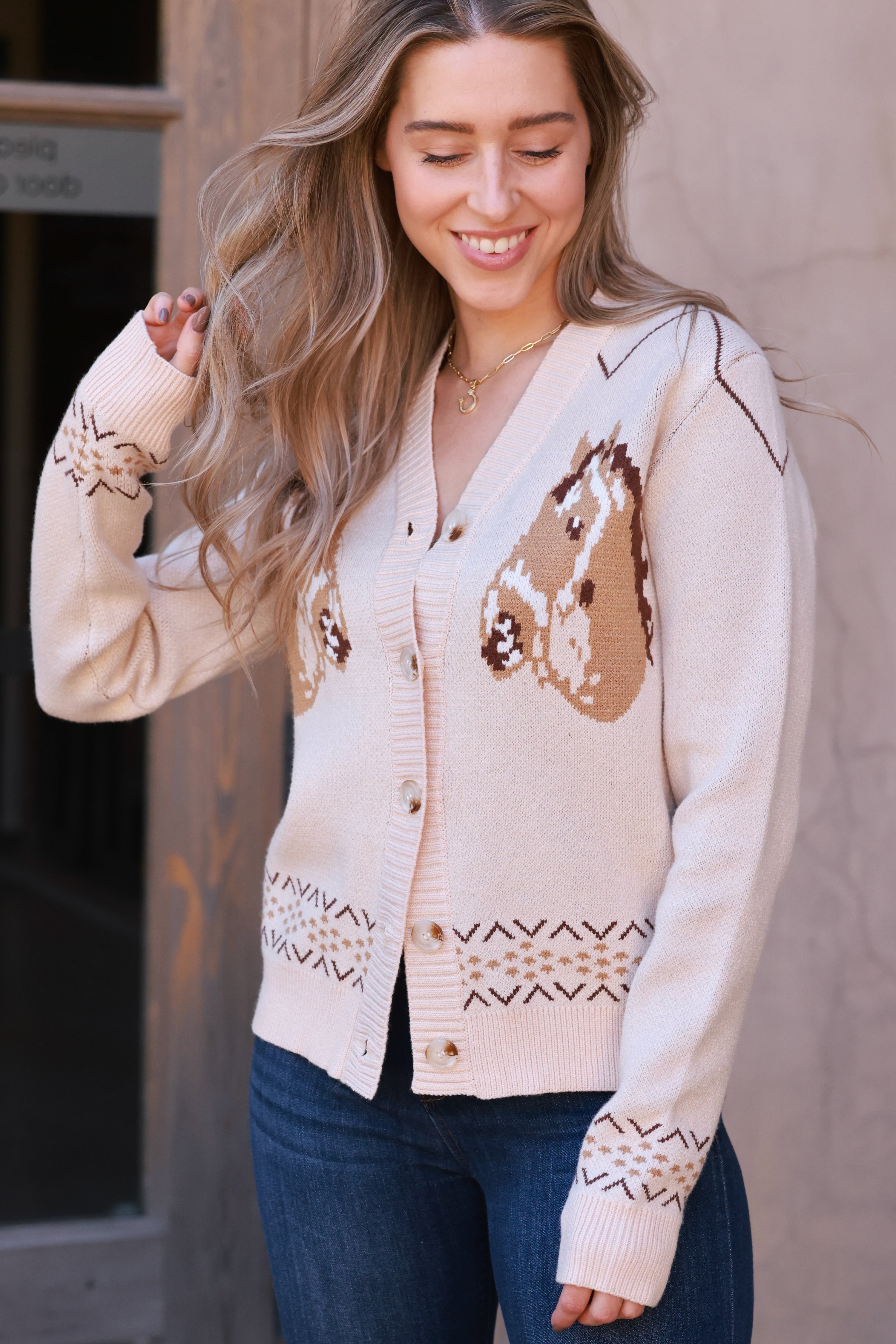 A cozy cream-colored knit cardigan with two brown horse graphics and subtle geometric fair isle trim. This long-sleeve, V-neck button-down features a relaxed, slightly cropped fit and soft viscose-poly blend fabric, offering a playful, equestrian-inspired look perfect for fall.