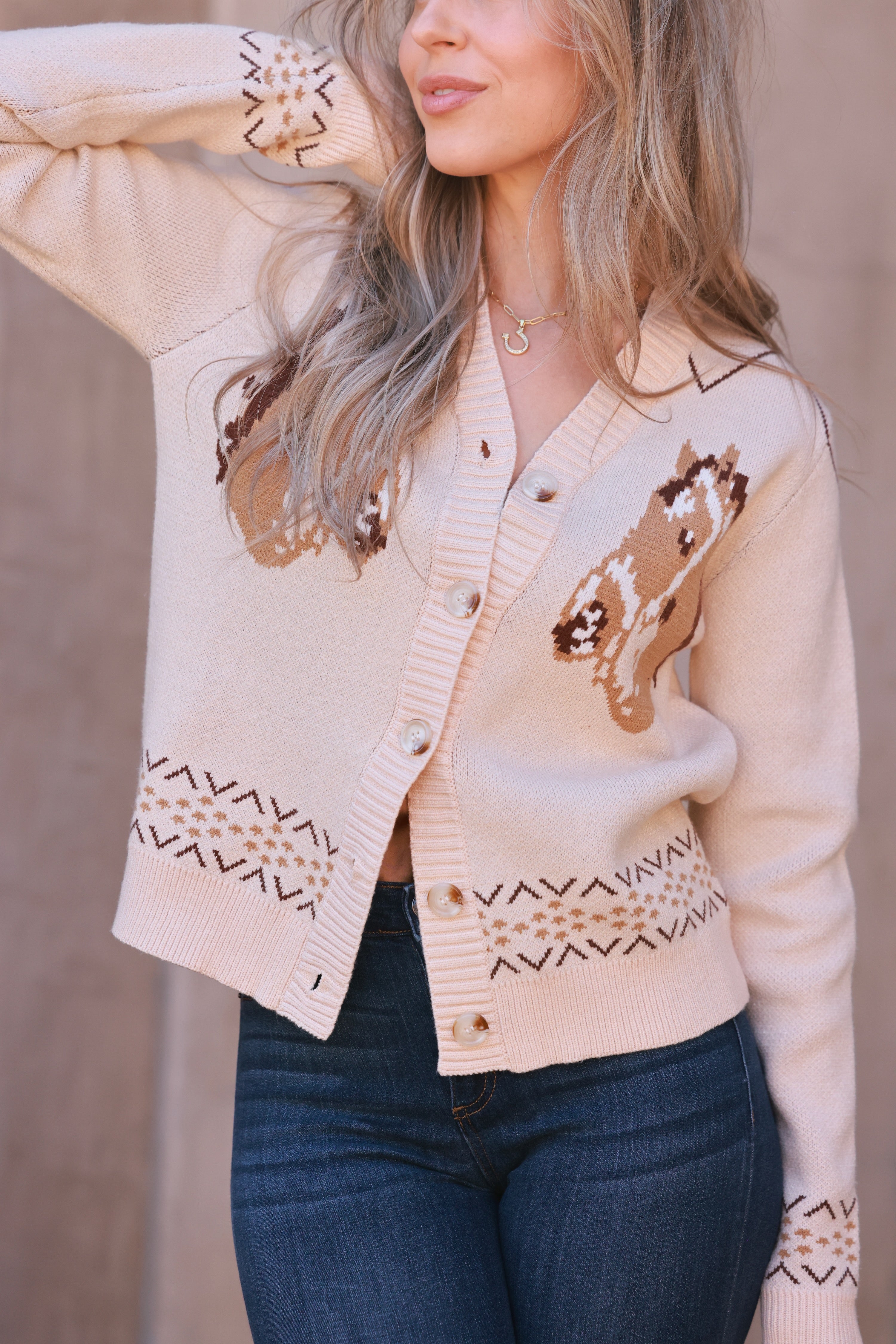 A cozy cream-colored knit cardigan with two brown horse graphics and subtle geometric fair isle trim. This long-sleeve, V-neck button-down features a relaxed, slightly cropped fit and soft viscose-poly blend fabric, offering a playful, equestrian-inspired look perfect for fall.