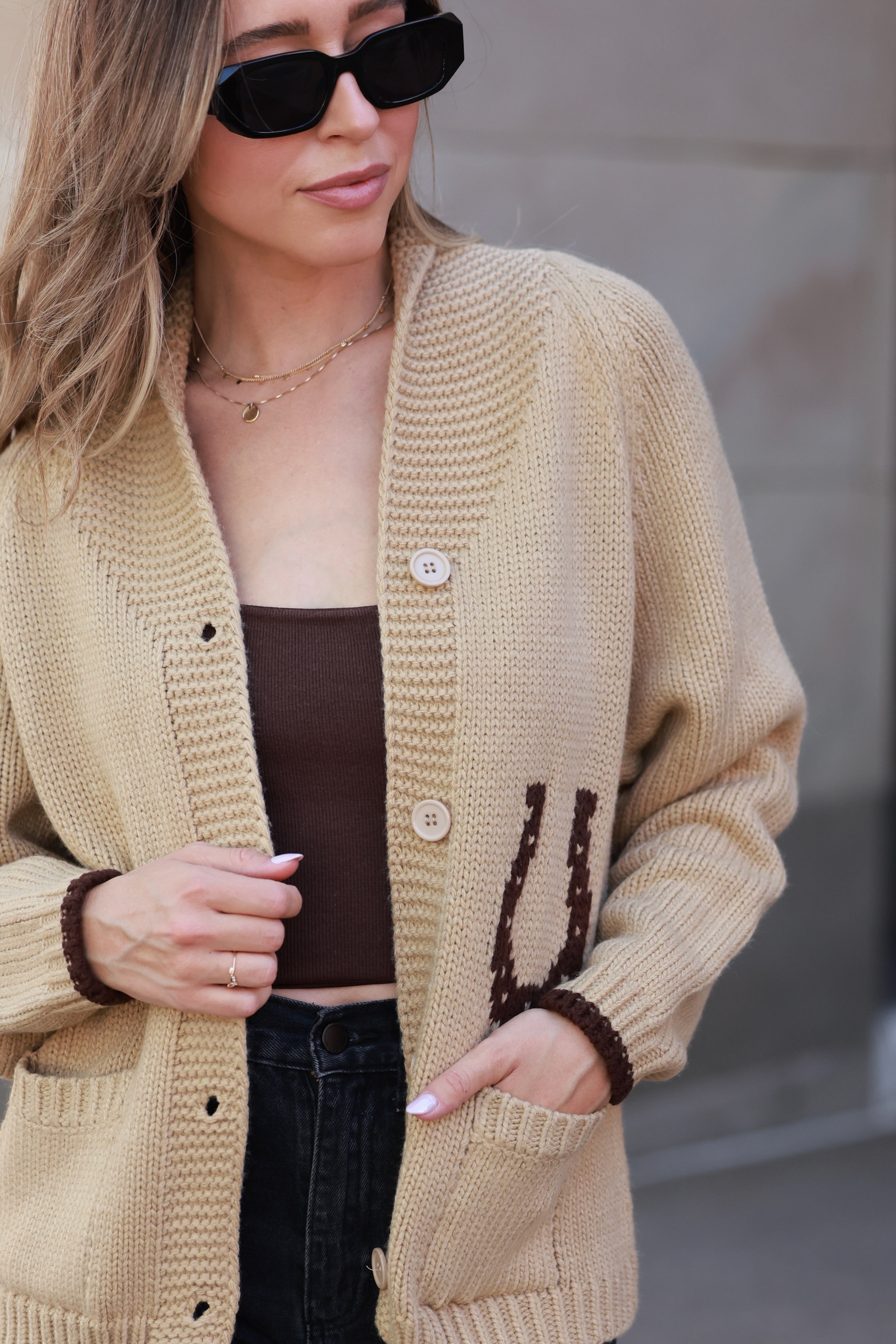 Woman wearing a beige cardigan with brown trim, sunglasses, and a blurred background - Varsity cardigan, Horse graphic sweater, Equestrian knitwear, Western statement cardigan, Camel horse cardigan